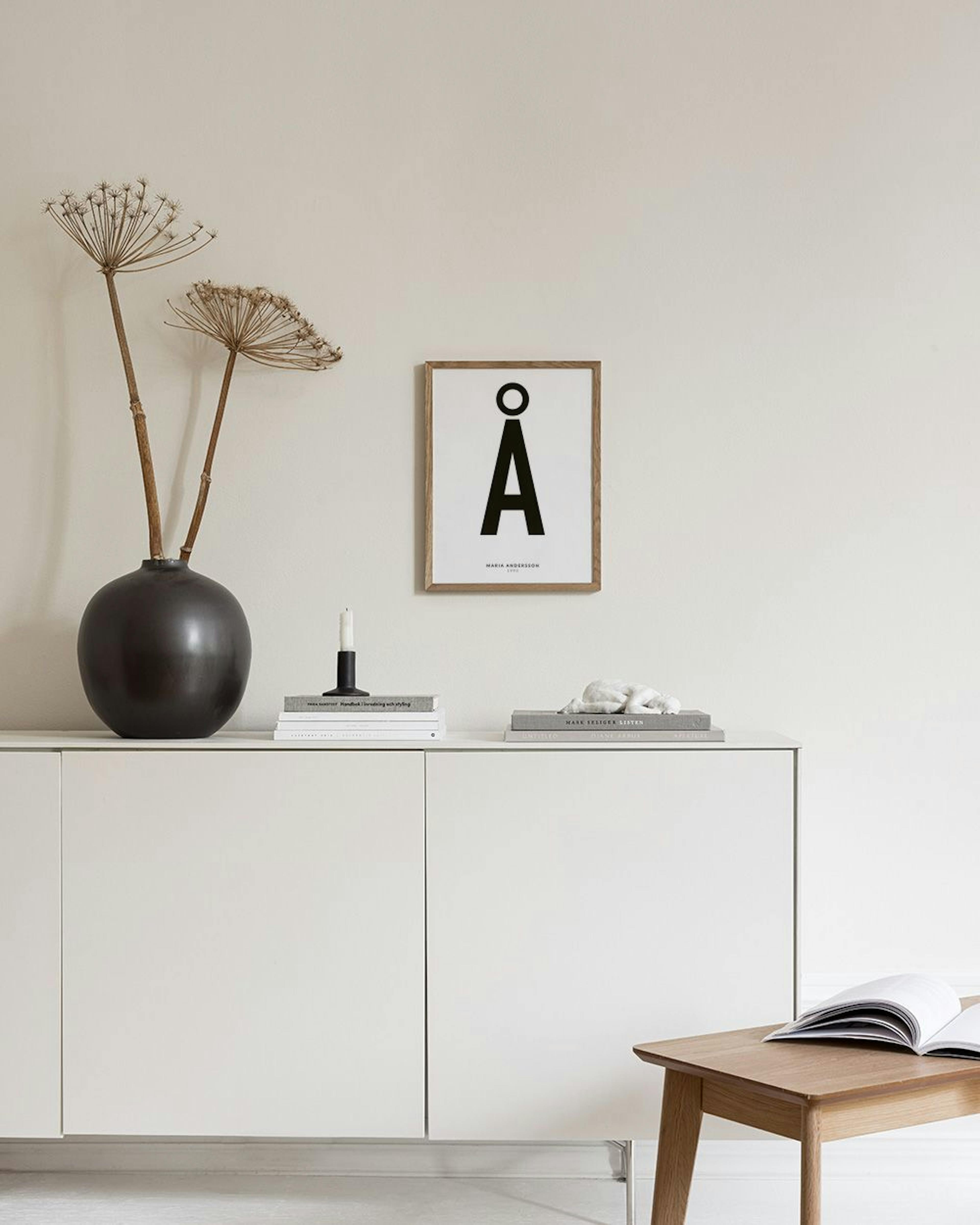 Poster personalizado com um grande Å a preto, pendurado numa parede cinzenta acima de um móvel verde com livros e objetos decora