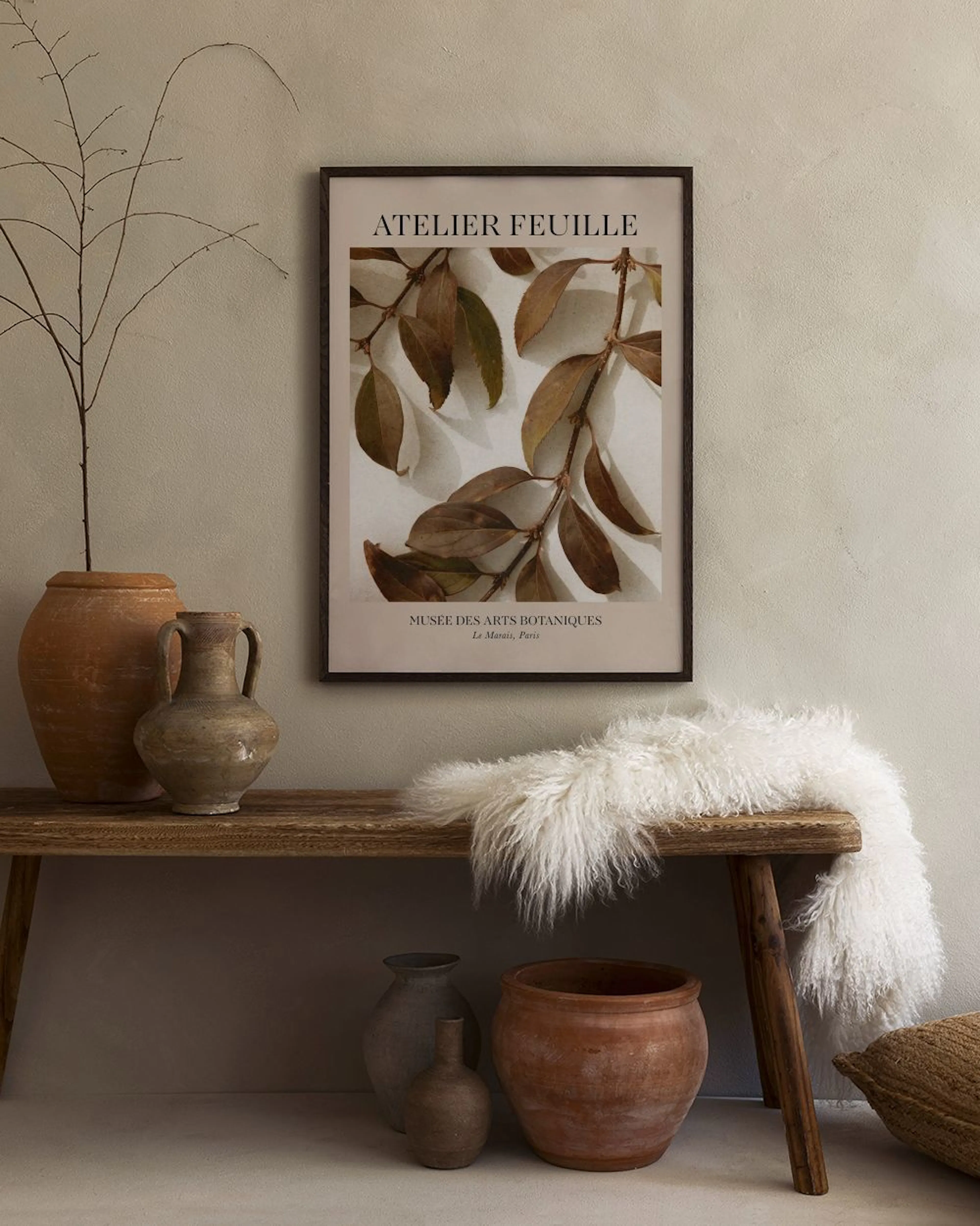 A botanical print of brown leaves on a light background, hanging above a wooden bench with rustic pots and a white sheepskin thr