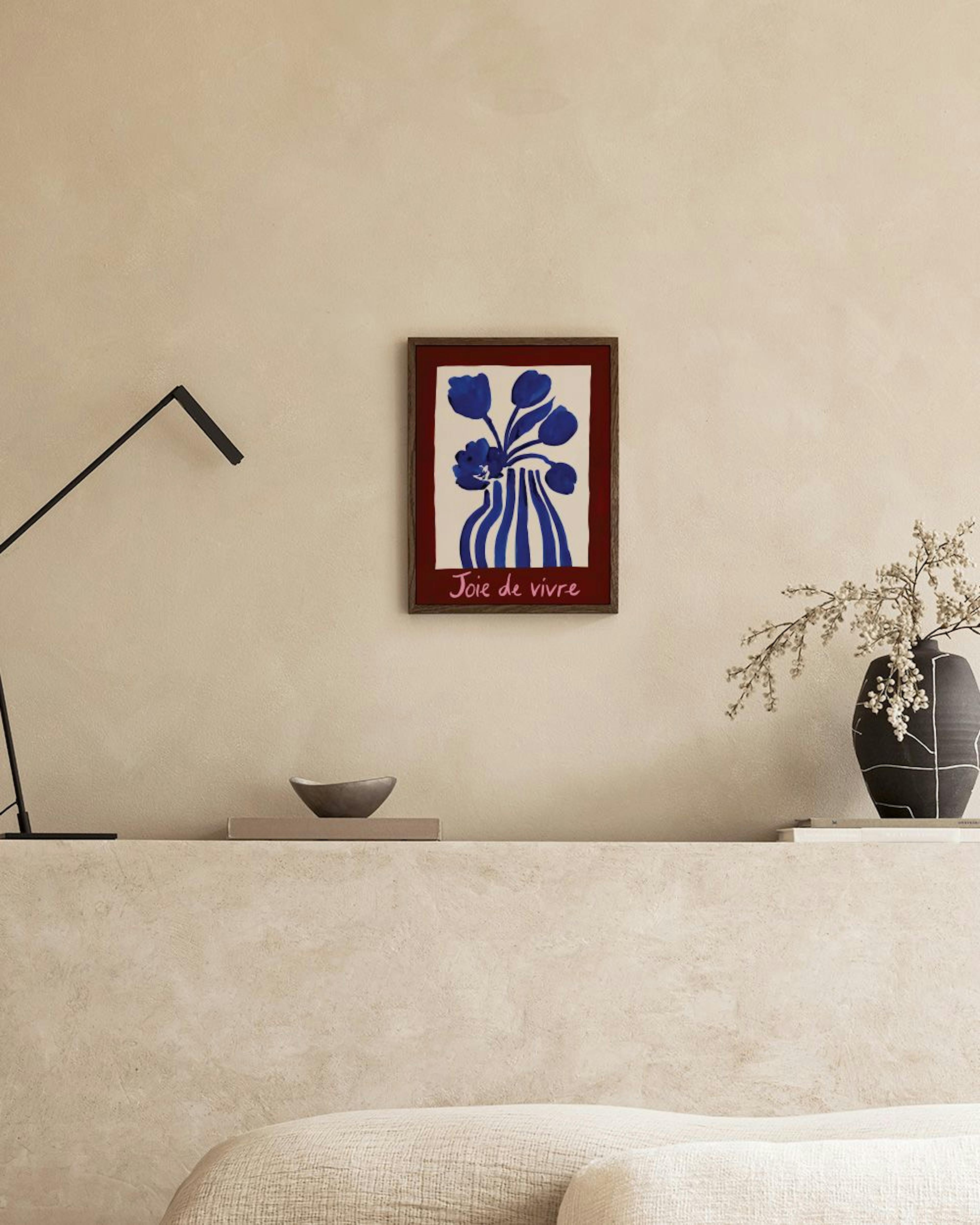 Plakat z niebieskimi kwiatami w paski i napisem Joie de vivre, zawieszony nad beżową sofą.