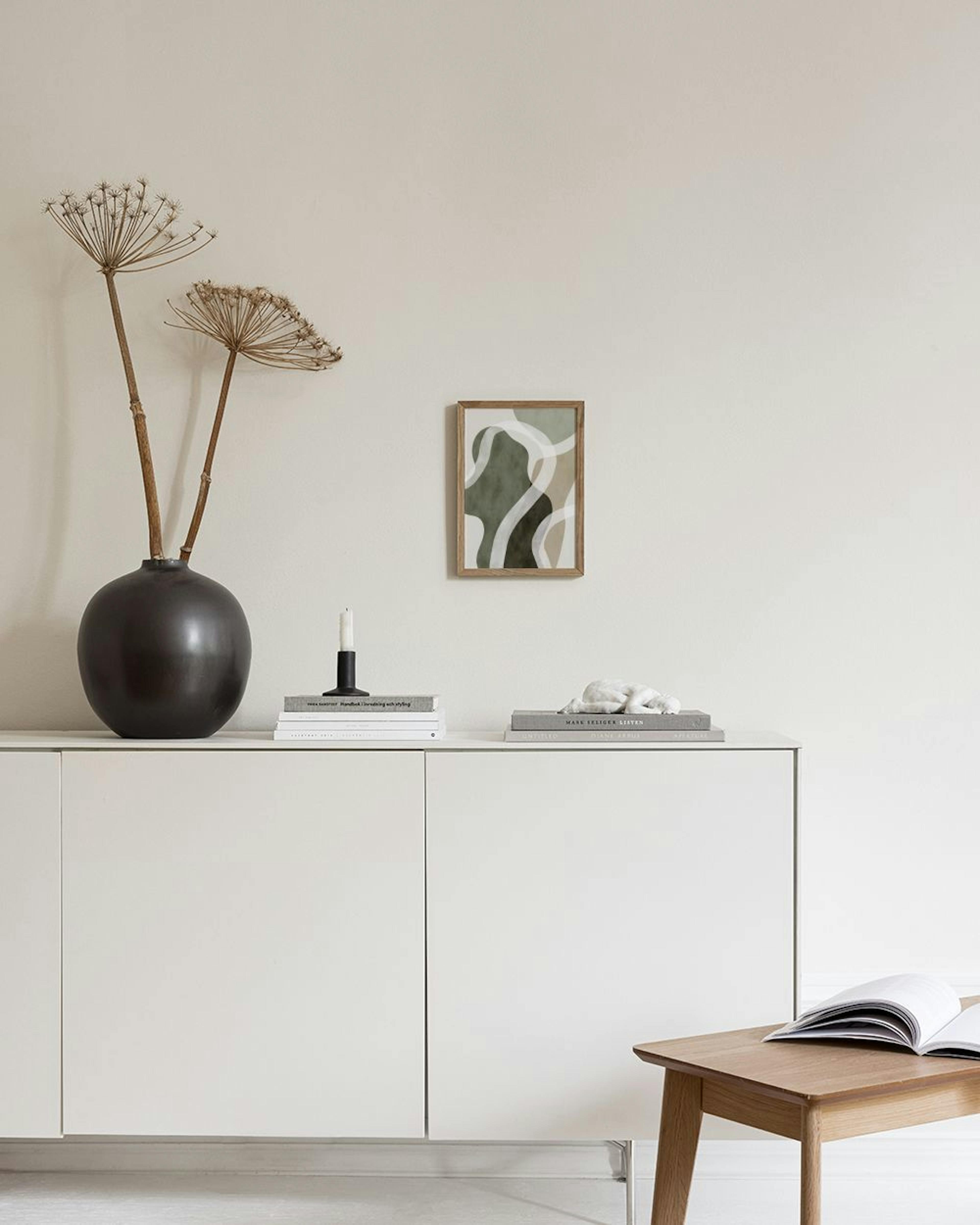 Poster mit organischen Formen in Grün-, Beige- und Brauntönen mit weißen Linien, über einem Hocker im Wohnzimmer.
