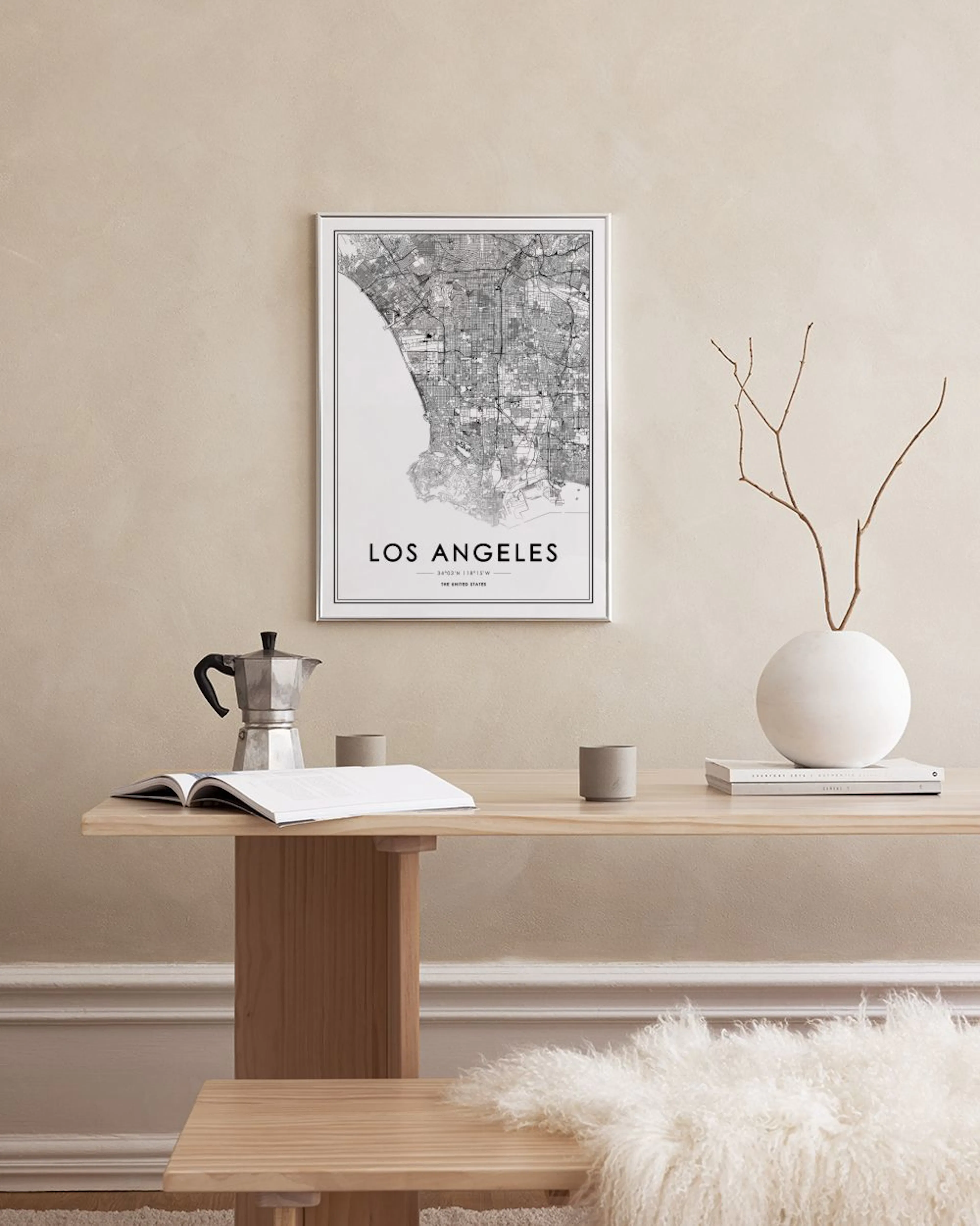 A Los Angeles map print hanging on a black grid, displayed in a room with a light wood chair, plant, and decorative vases.