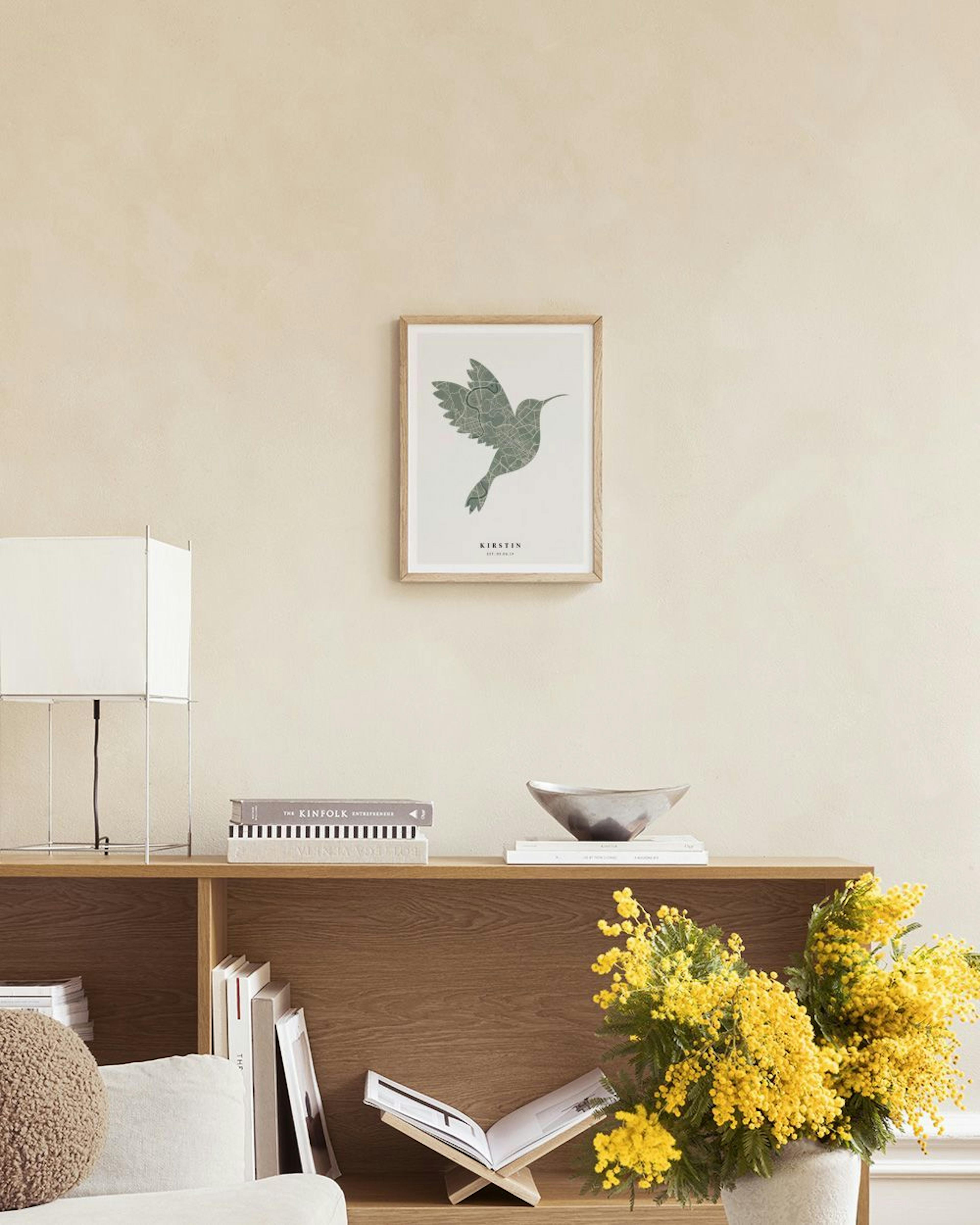Personalisiertes Poster mit Kolibri-Stadtplan, Name KIRSTIN und Datum, über einem grauen Sessel in einem modernen Wohnzimmer.