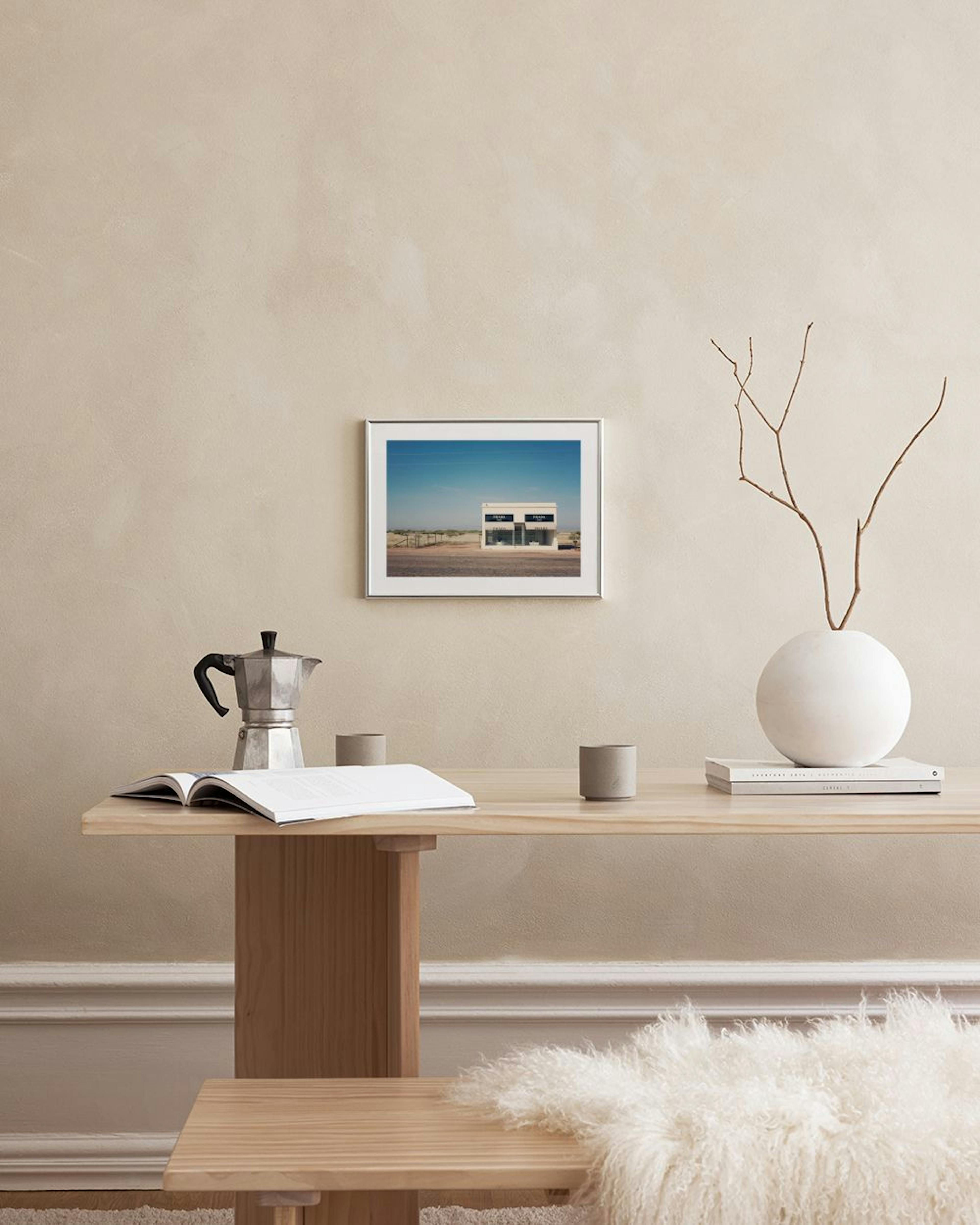 A print of a lone building with large windows in a desert landscape, hanging above a brown leather chair and white display stand