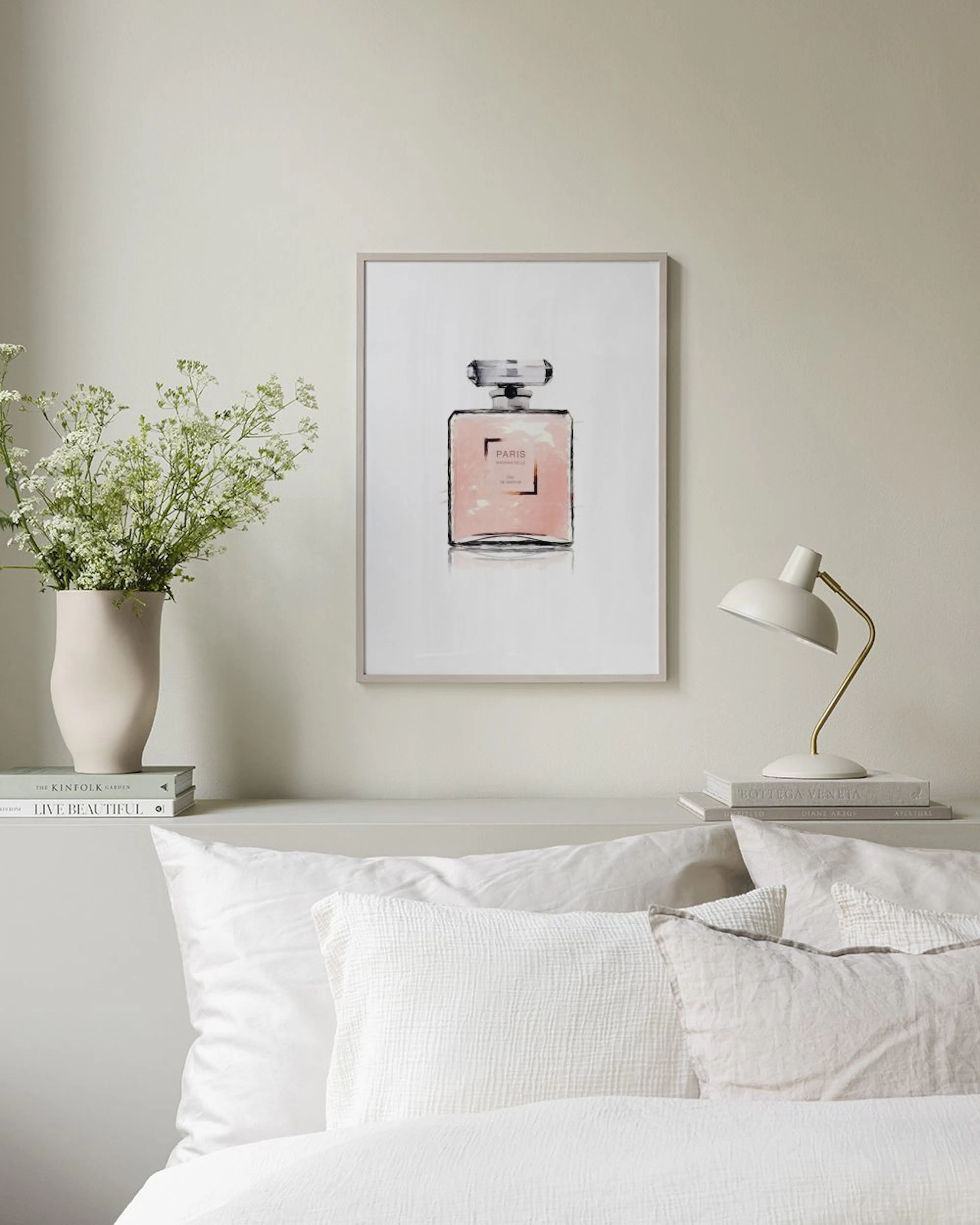Póster de una botella de perfume rosa estilo acuarela con PARIO en el centro, colgado en una habitación moderna. Se ven un silló