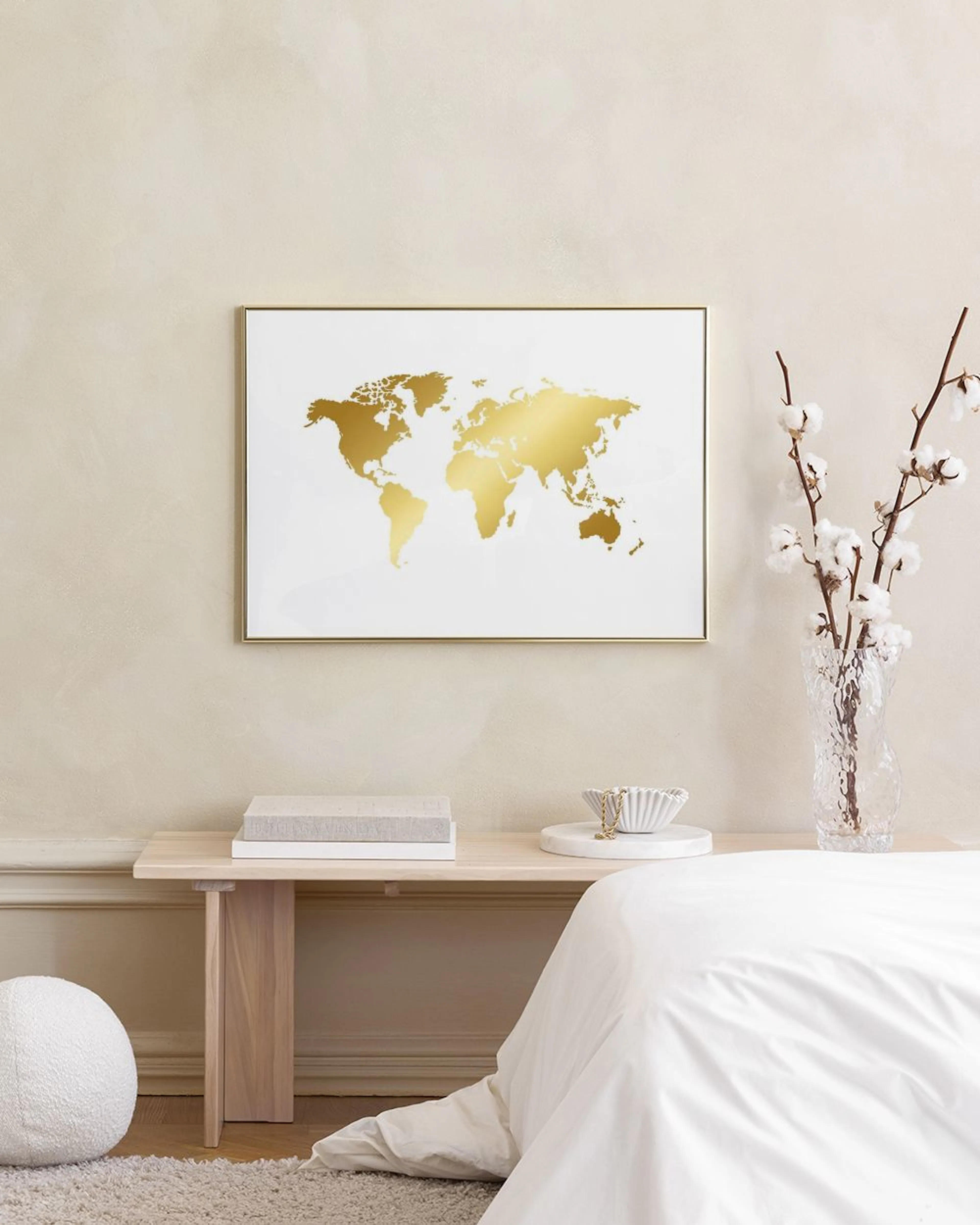 Póster de un mapa del mundo dorado en fondo blanco, colgando sobre una mesa auxiliar negra con libros y decoración, junto a un c