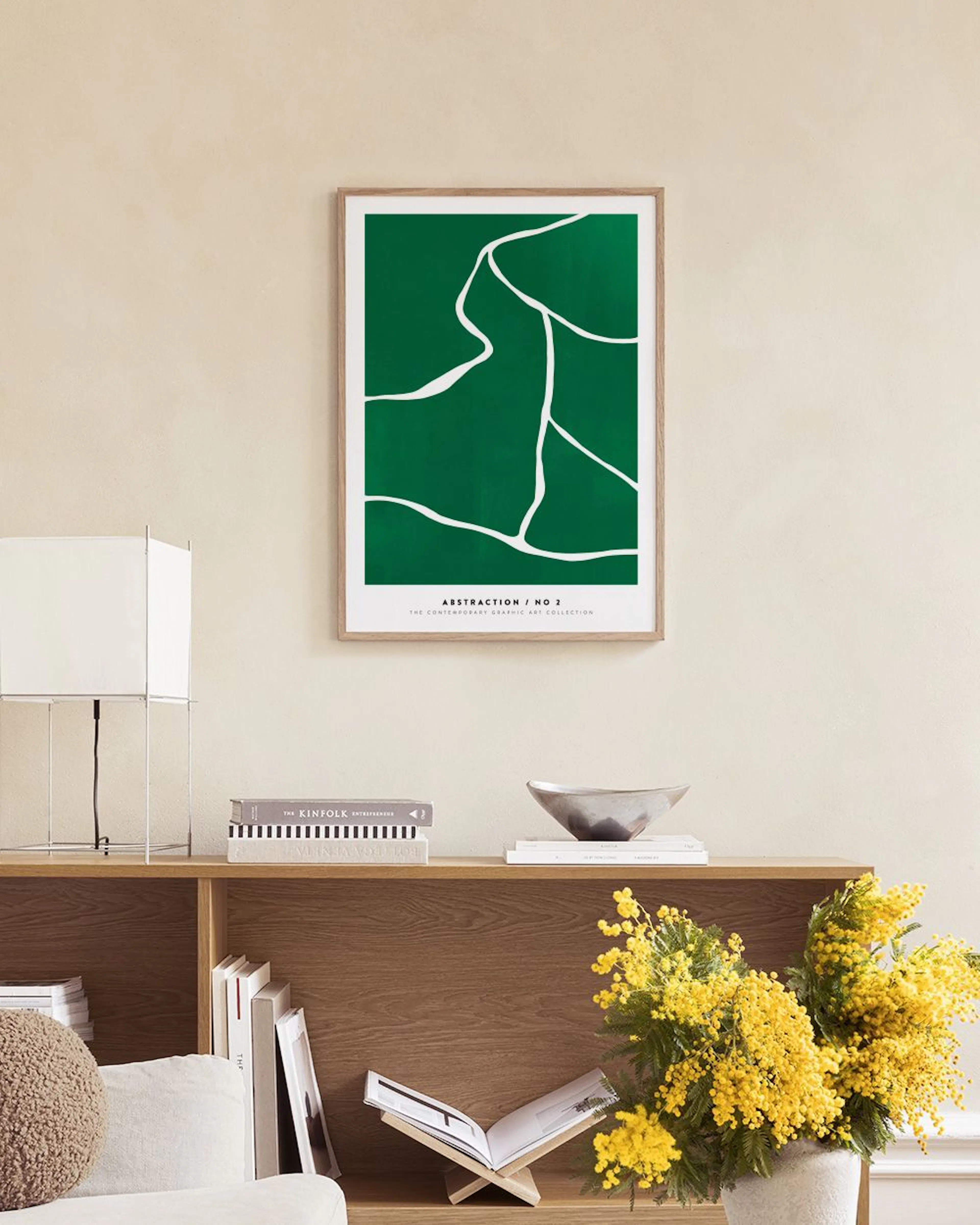 Póster abstracto verde con líneas blancas orgánicas colgado en una pared blanca, junto a una mesa redonda y un puf verde en una 