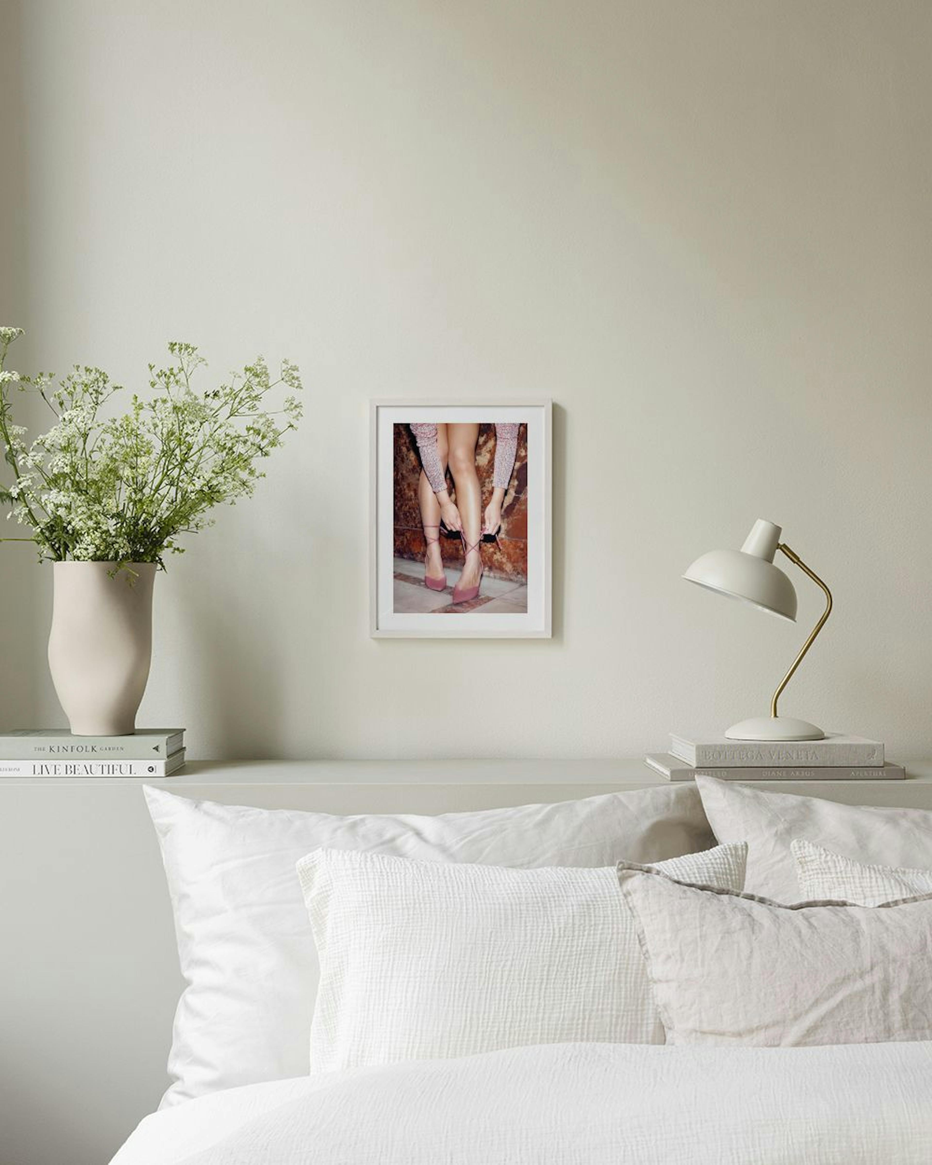 Poster con gambe femminili che allacciano tacchi rosa, appeso sopra una panca rosa in un ambiente moderno.