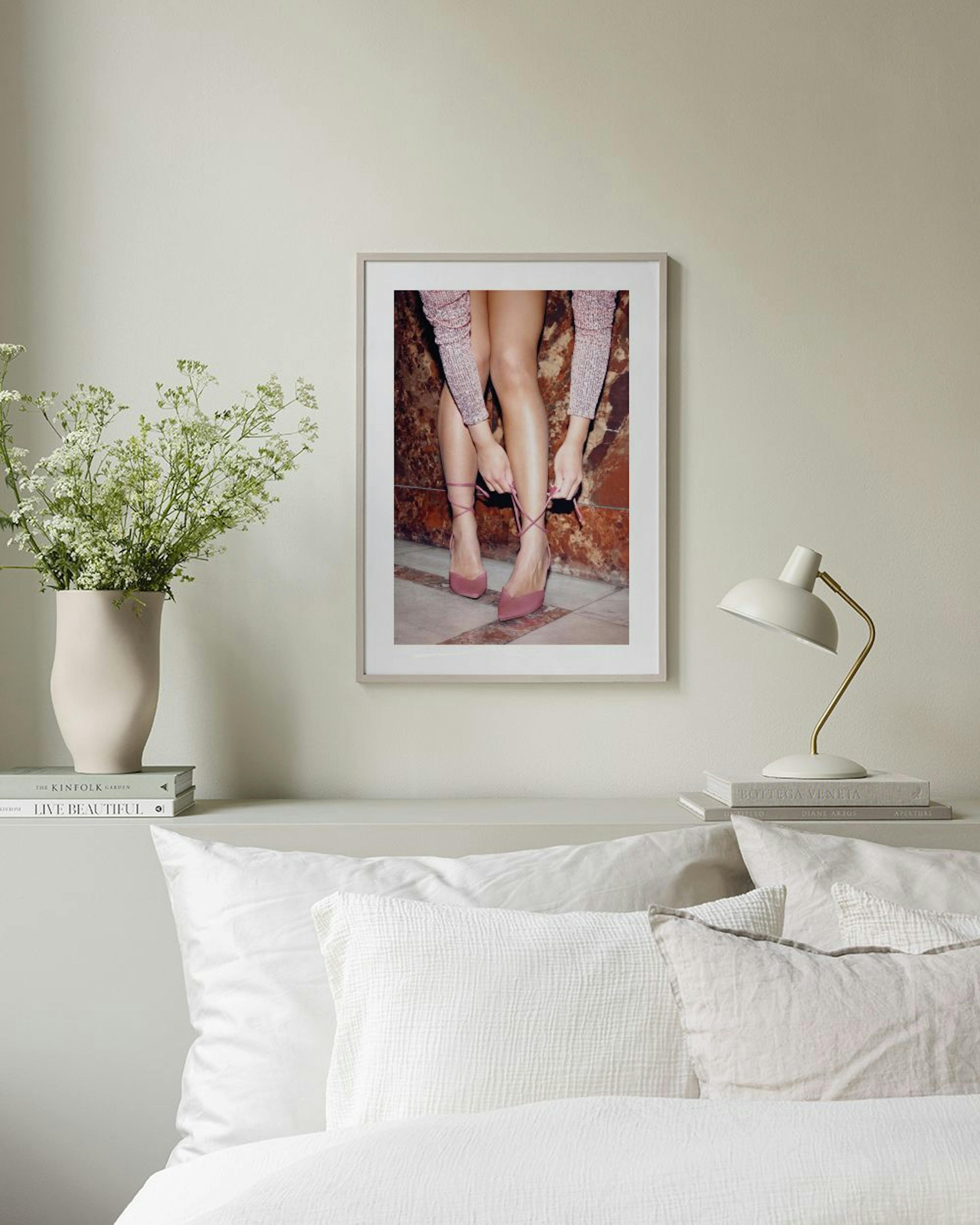 A fashion print of a womans legs tying pink lace-up heels, hanging above a pink velvet bench in a room.