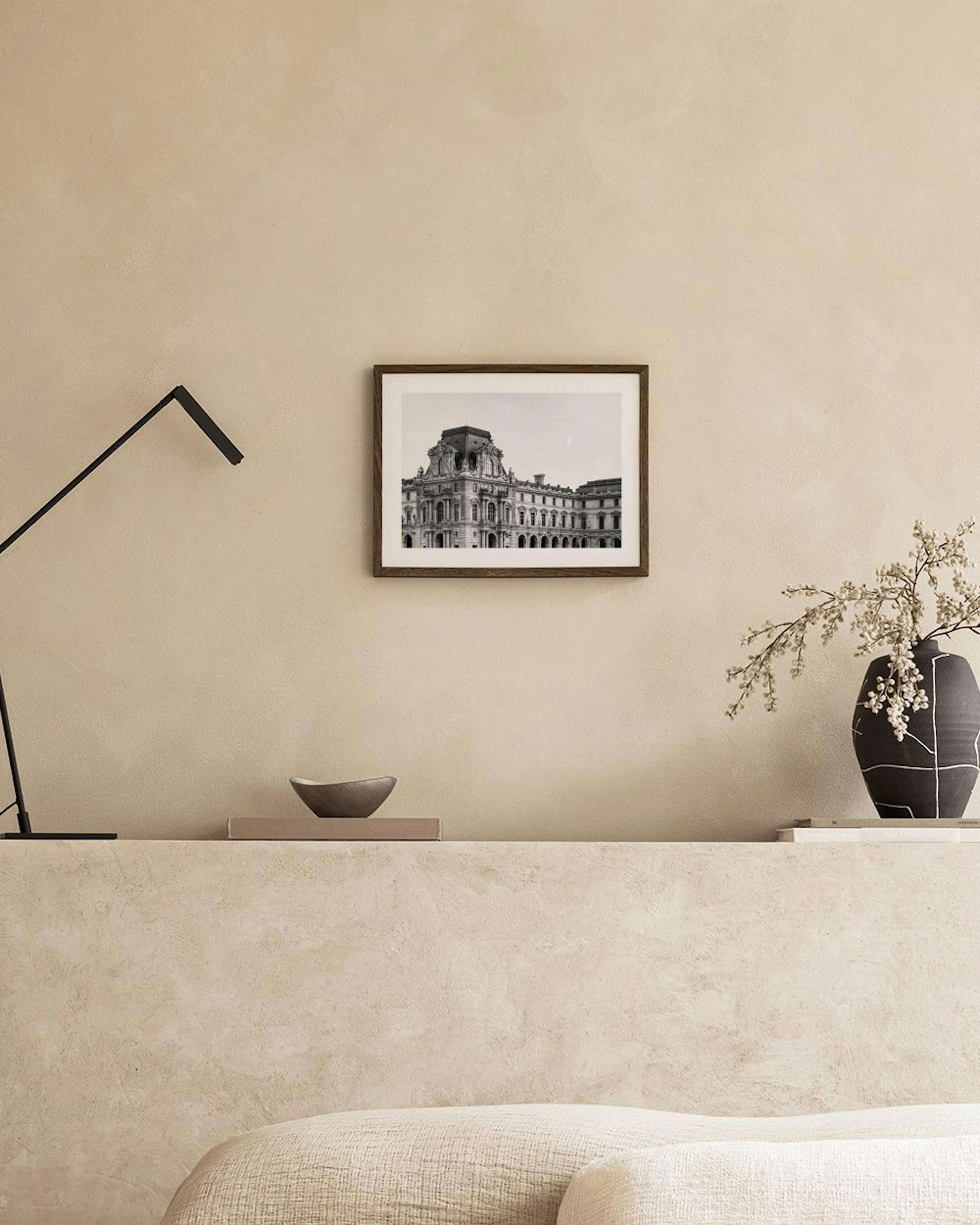 Poster in bianco e nero del Palazzo del Louvre, appeso sopra una credenza in rattan con ciotola e libri.