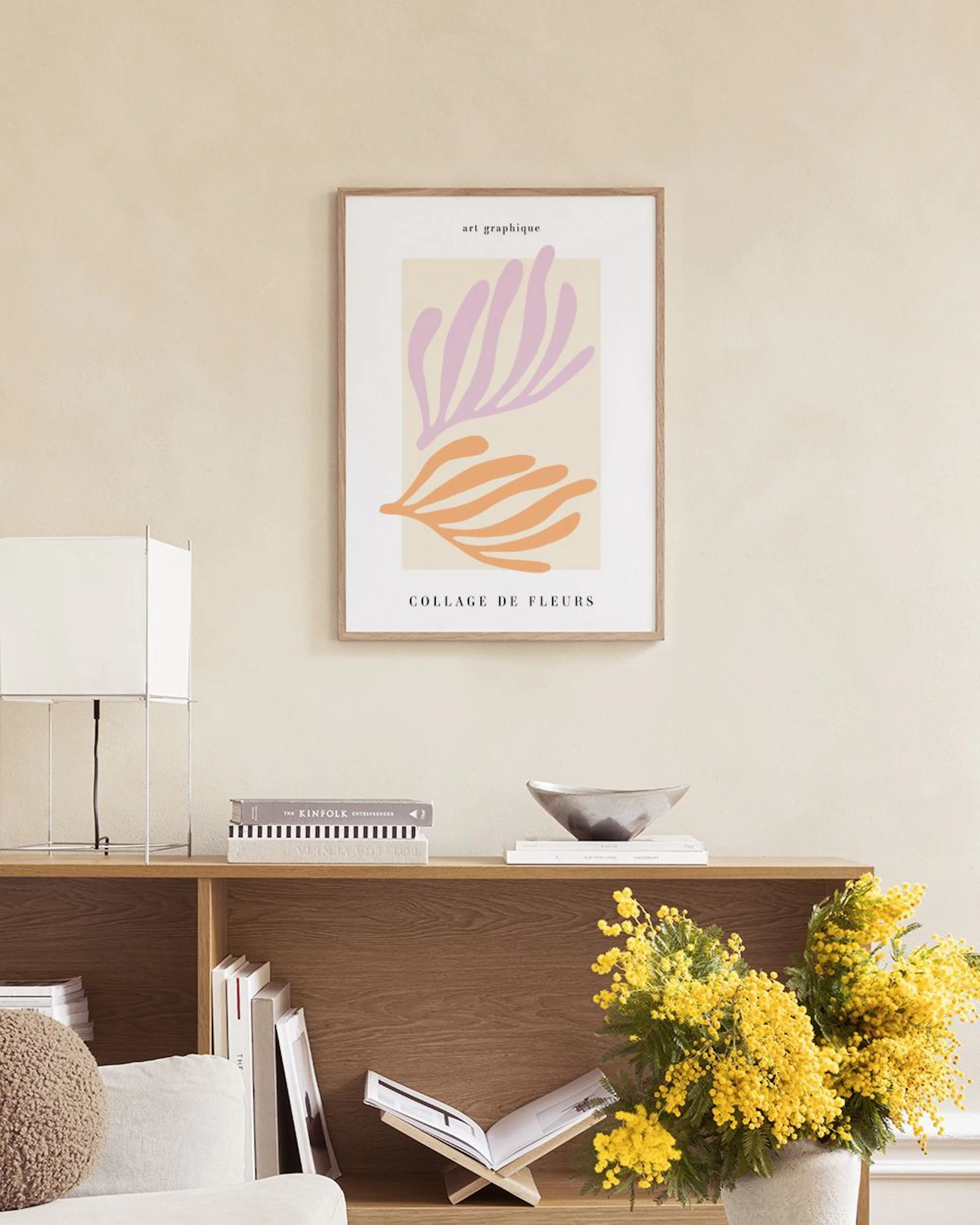 Poster con forme astratte lilla e arancioni su sfondo chiaro, appeso sopra un tavolino bianco con vasi e libri.