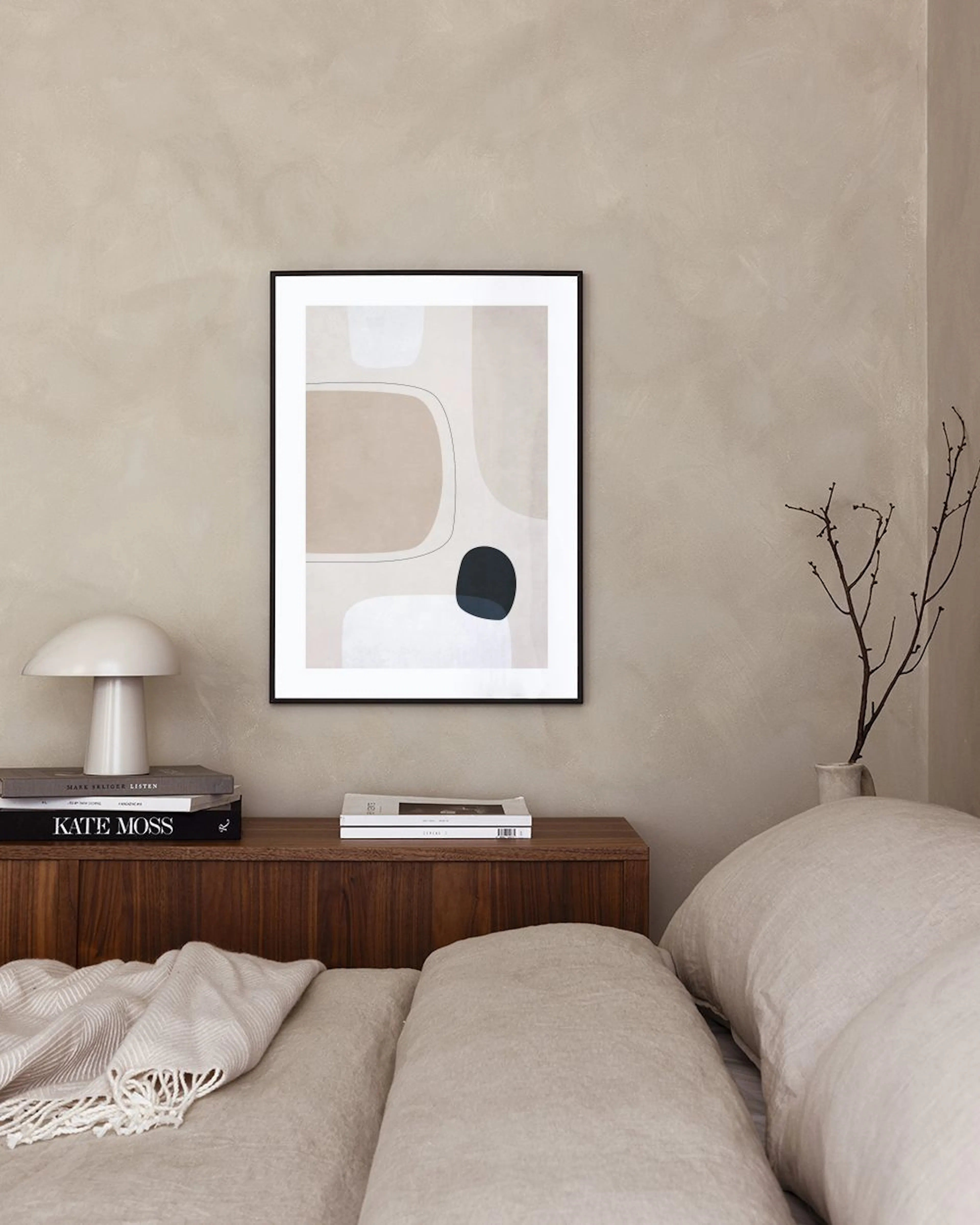 Poster med abstrakta former i beige och vitt med svarta detaljer, hängande i ett modernt rum med en rottingstol och en vas med t