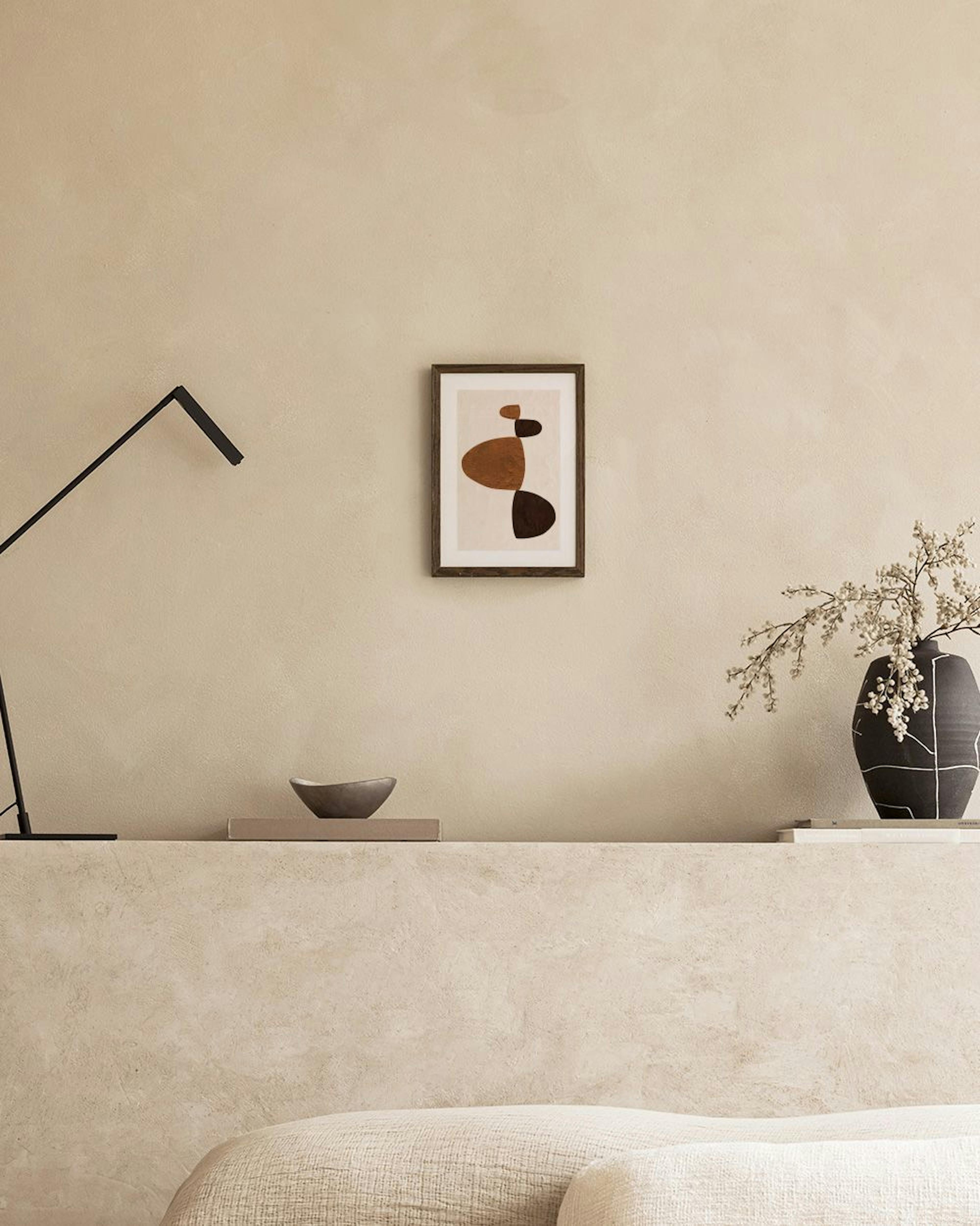 Poster com formas orgânicas em tons castanhos e creme, pendurado acima de uma mesa lateral com um vaso e livro.