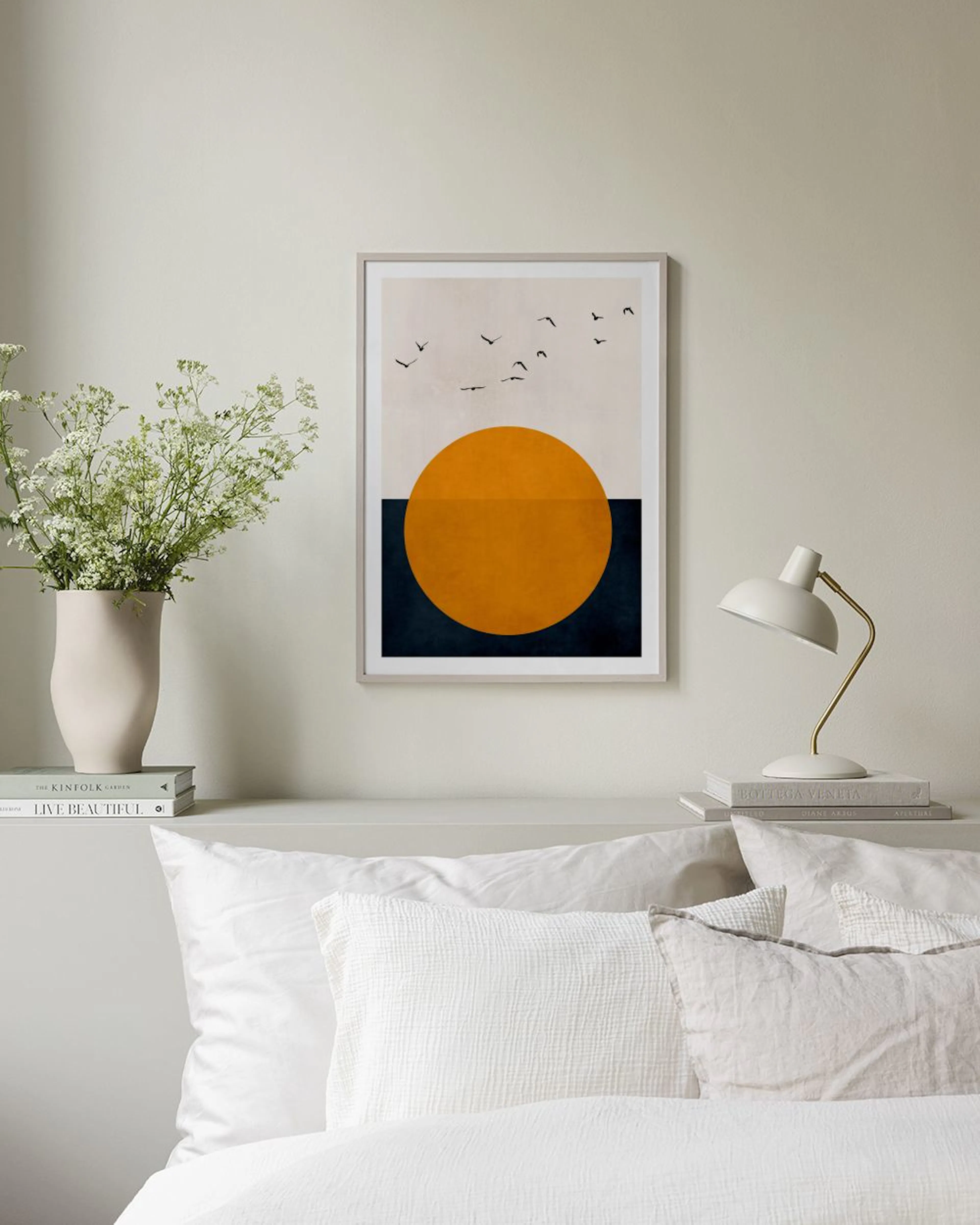 Poster met abstracte oranje zon en vliegende vogels, hangend aan een grijze muur boven een zwarte bijzettafel.