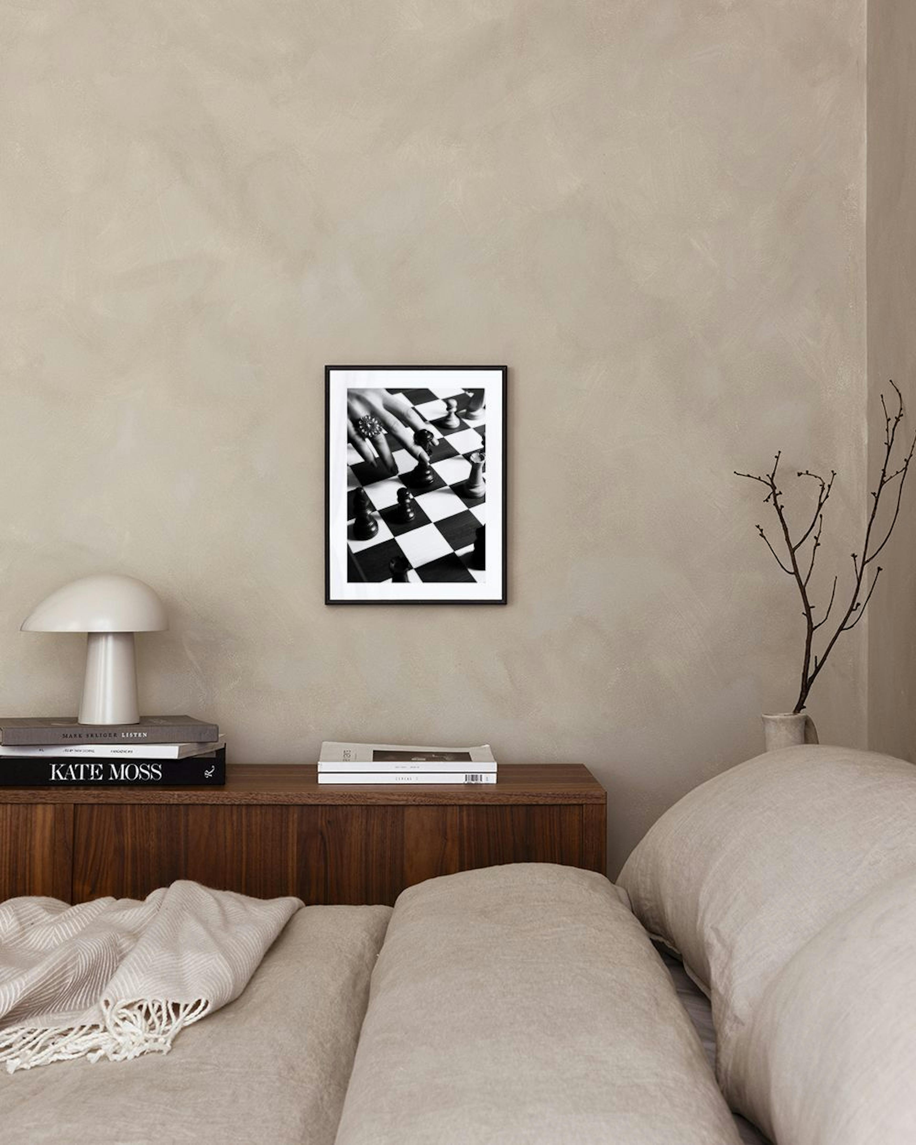 Black and white print of a hand with a ring moving a chess piece on a chessboard, displayed on a grey console table.