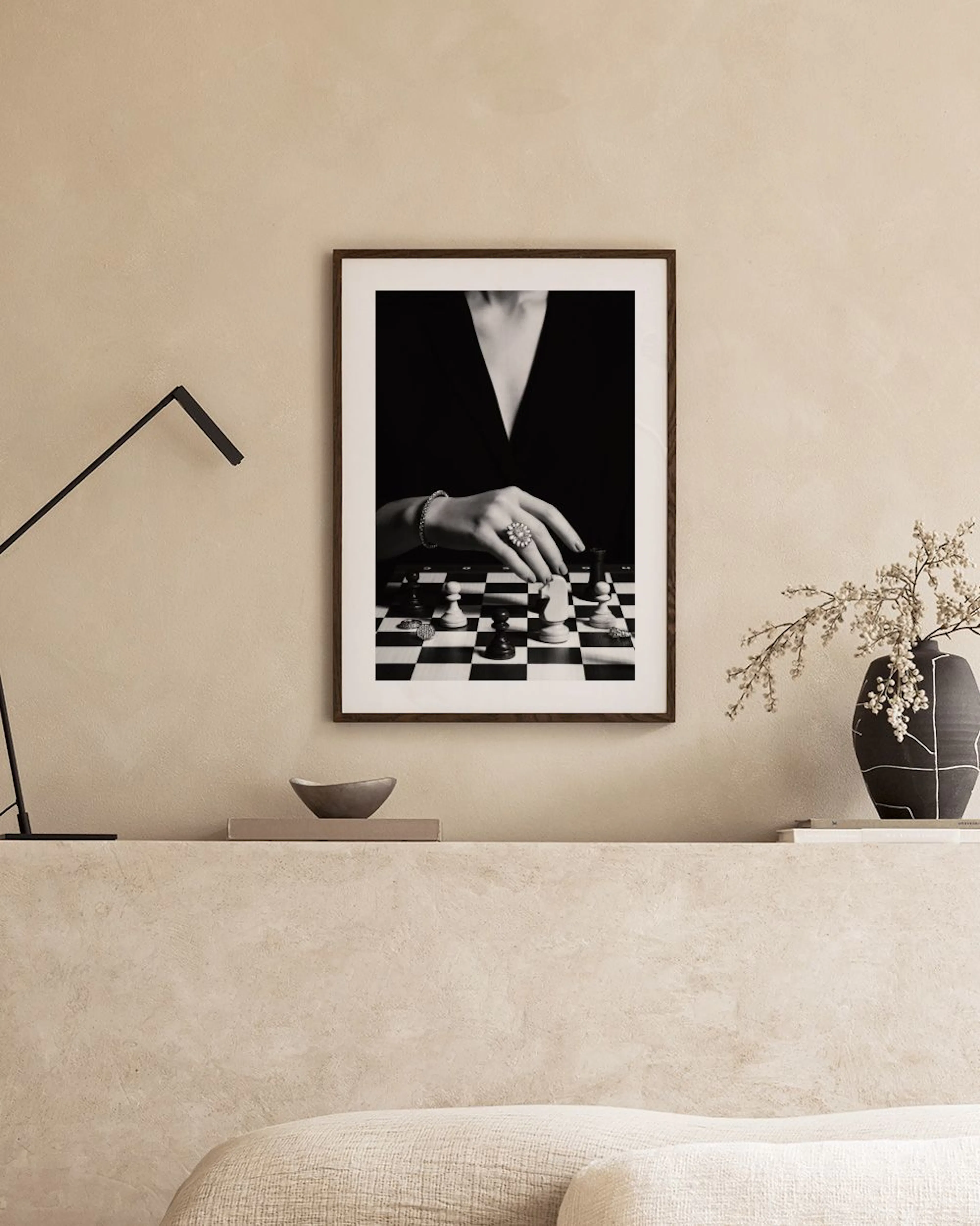 Póster en blanco y negro de una mano con anillo y pulsera moviendo una pieza de ajedrez, colgado en una pared clara.