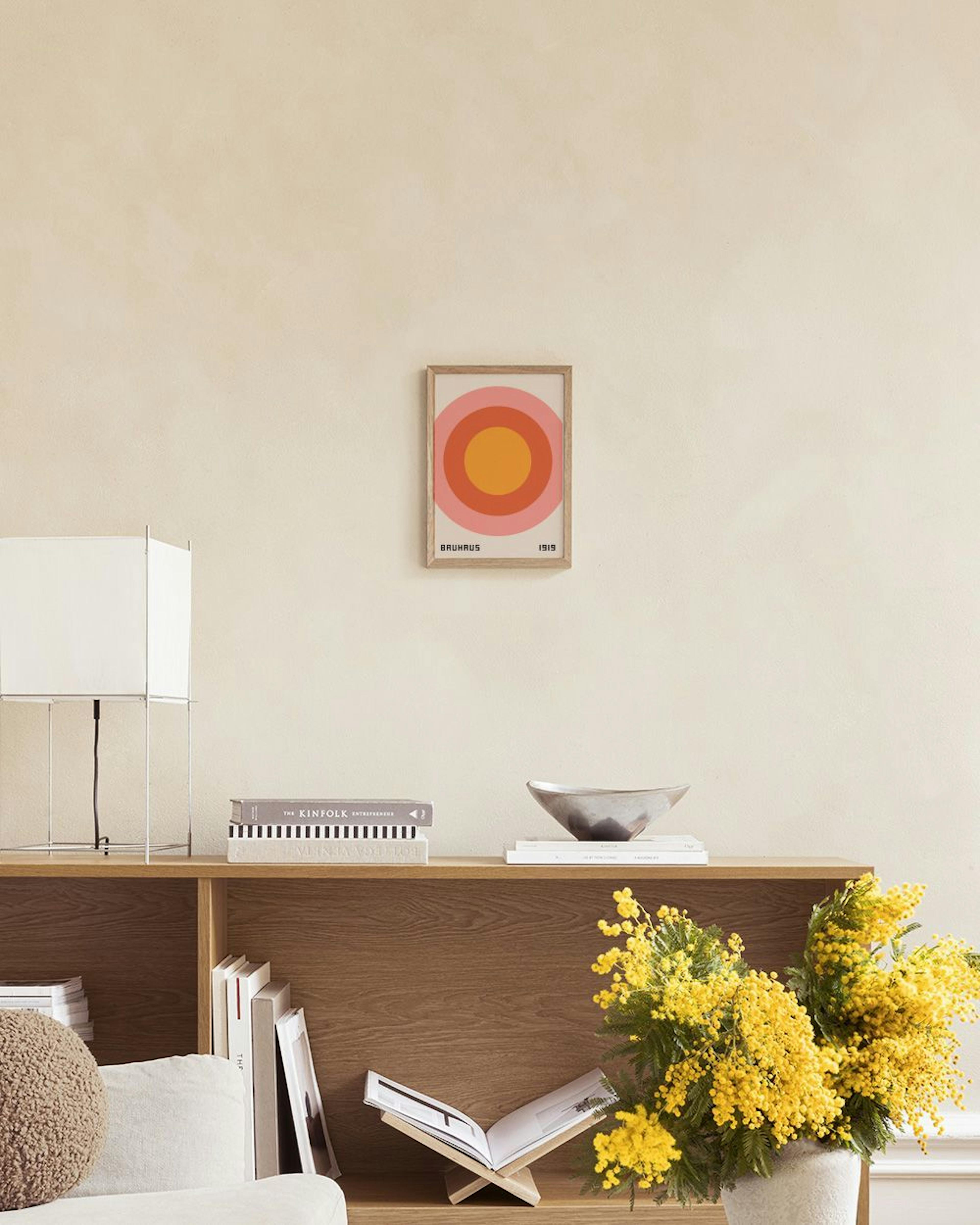 Poster met Bauhaus-stijl, concentrische cirkels in oranje en roze, hangend boven een houten dressoir met rietwerk.