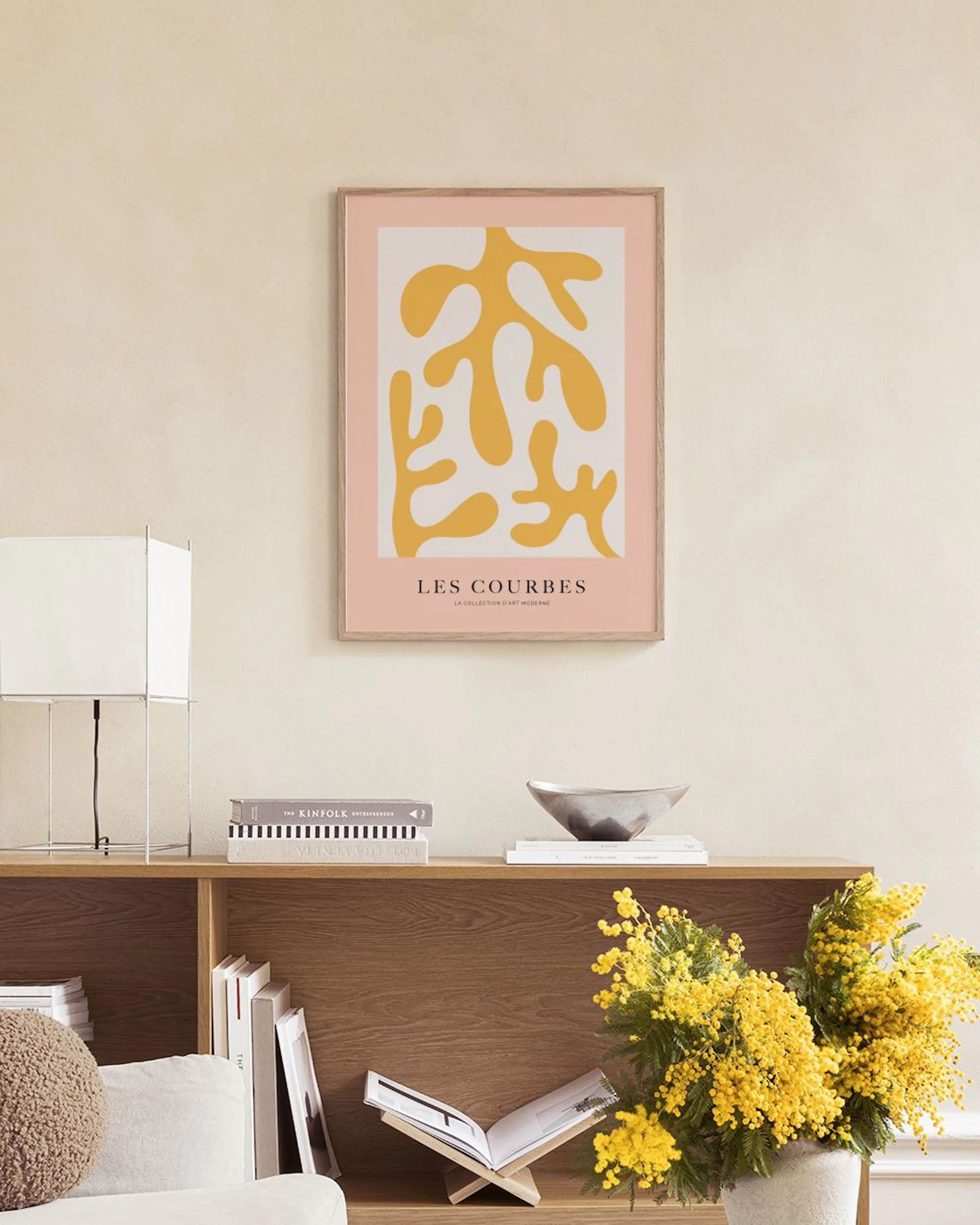 Poster mit gelben, organischen Formen auf weißem Grund und rosa Rand, hängt über einem Marmortisch mit Vasen und Büchern.