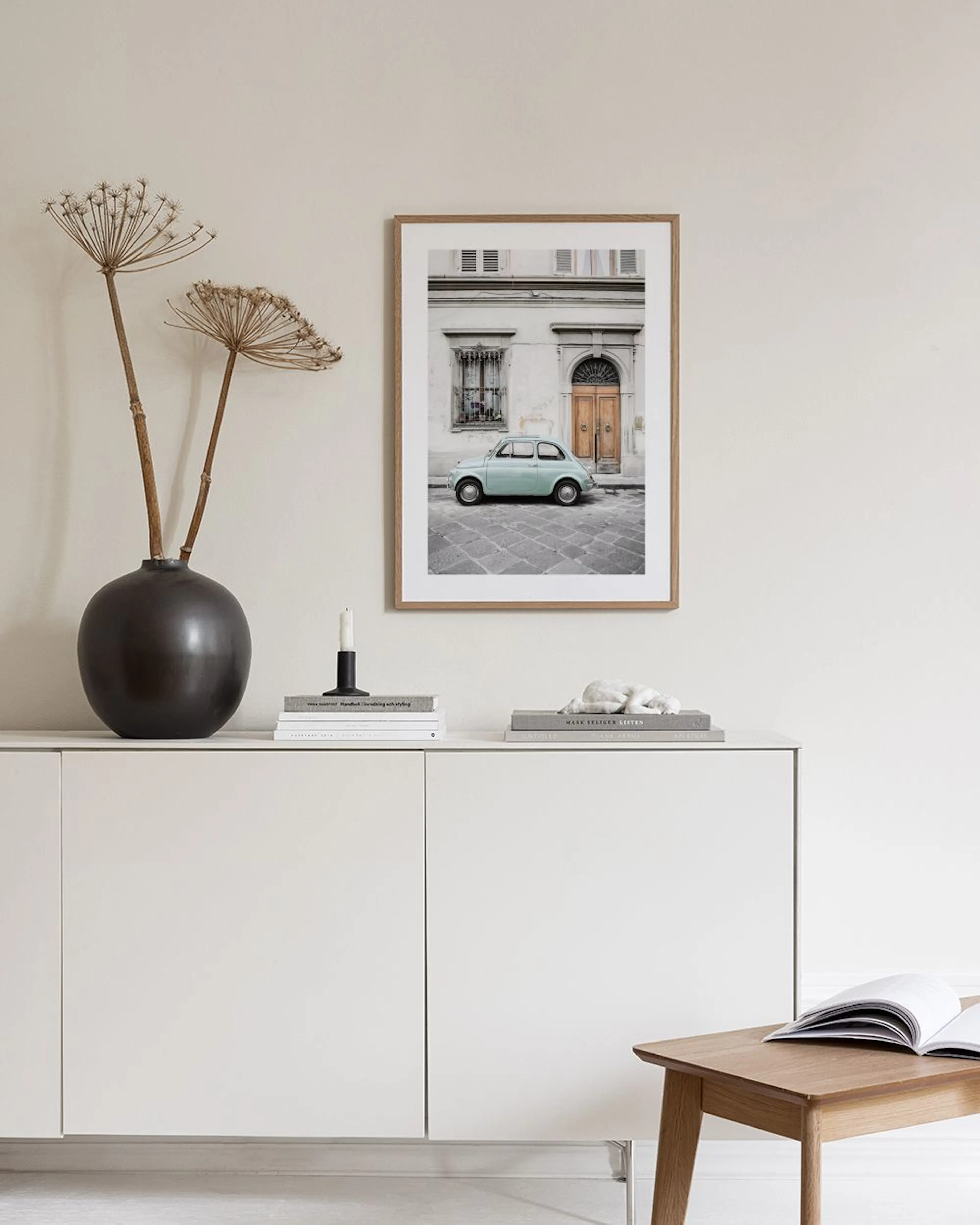 Plagát s mätovým autom Fiat 500 pred starou budovou, visí nad bielym nočným stolíkom s knihami a vázami.