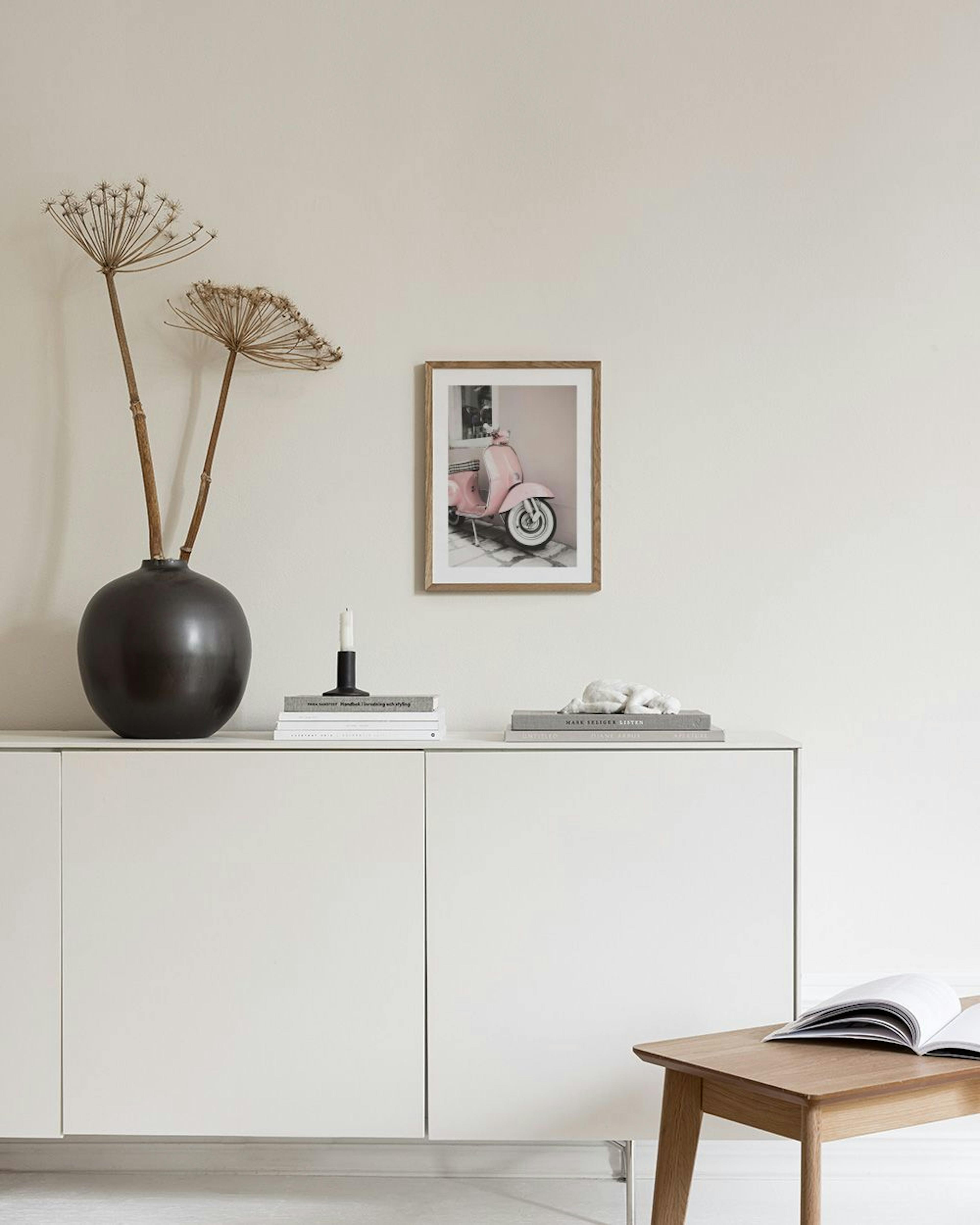 Poster van een roze Vespa scooter met witte banden, hangend aan een muur boven een roze poef en naast een vaas met bloemen.
