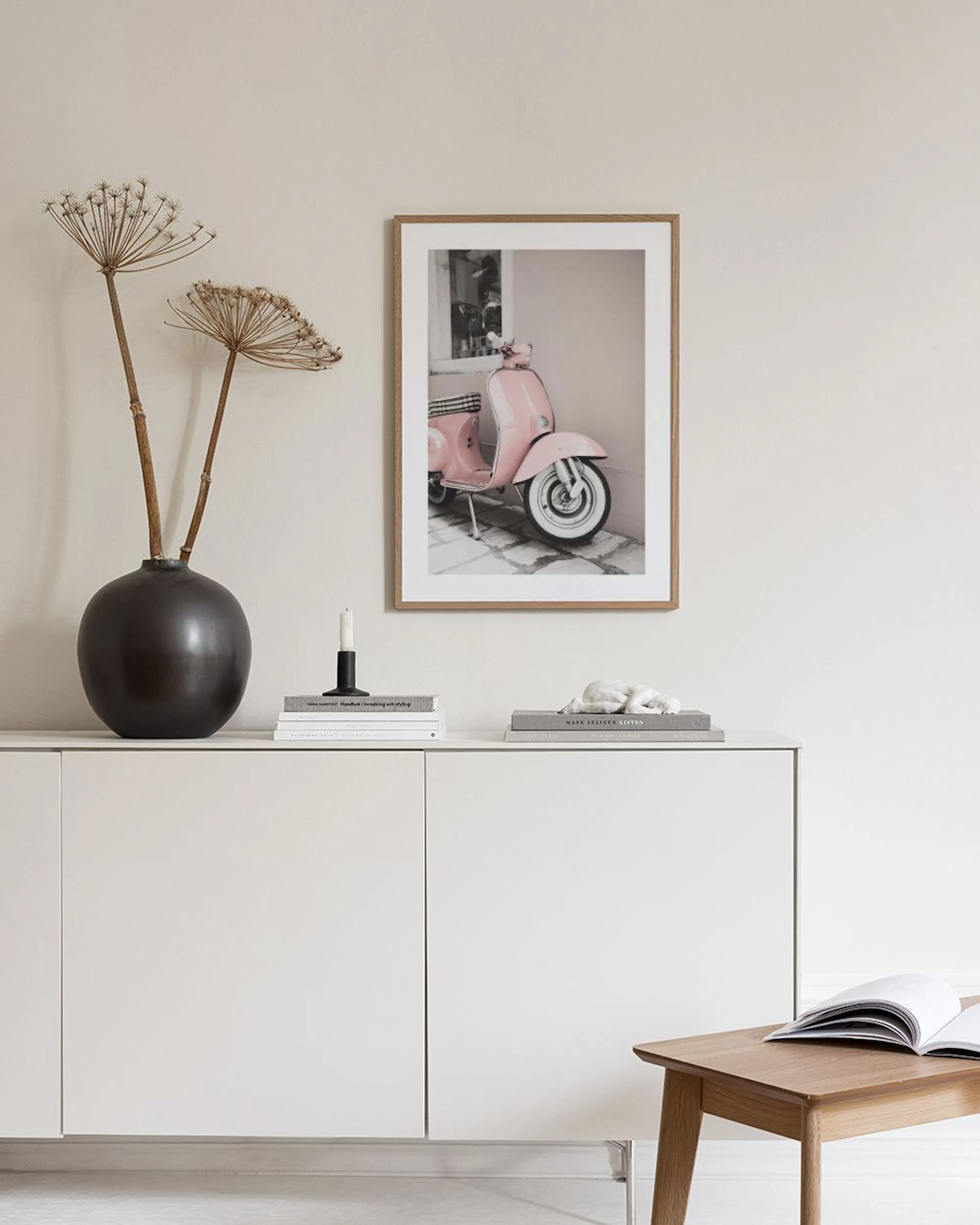 Print of a vintage pink scooter by a pale pink wall, displayed on a light gray wall with a vase of flowers and a pink ottoman.