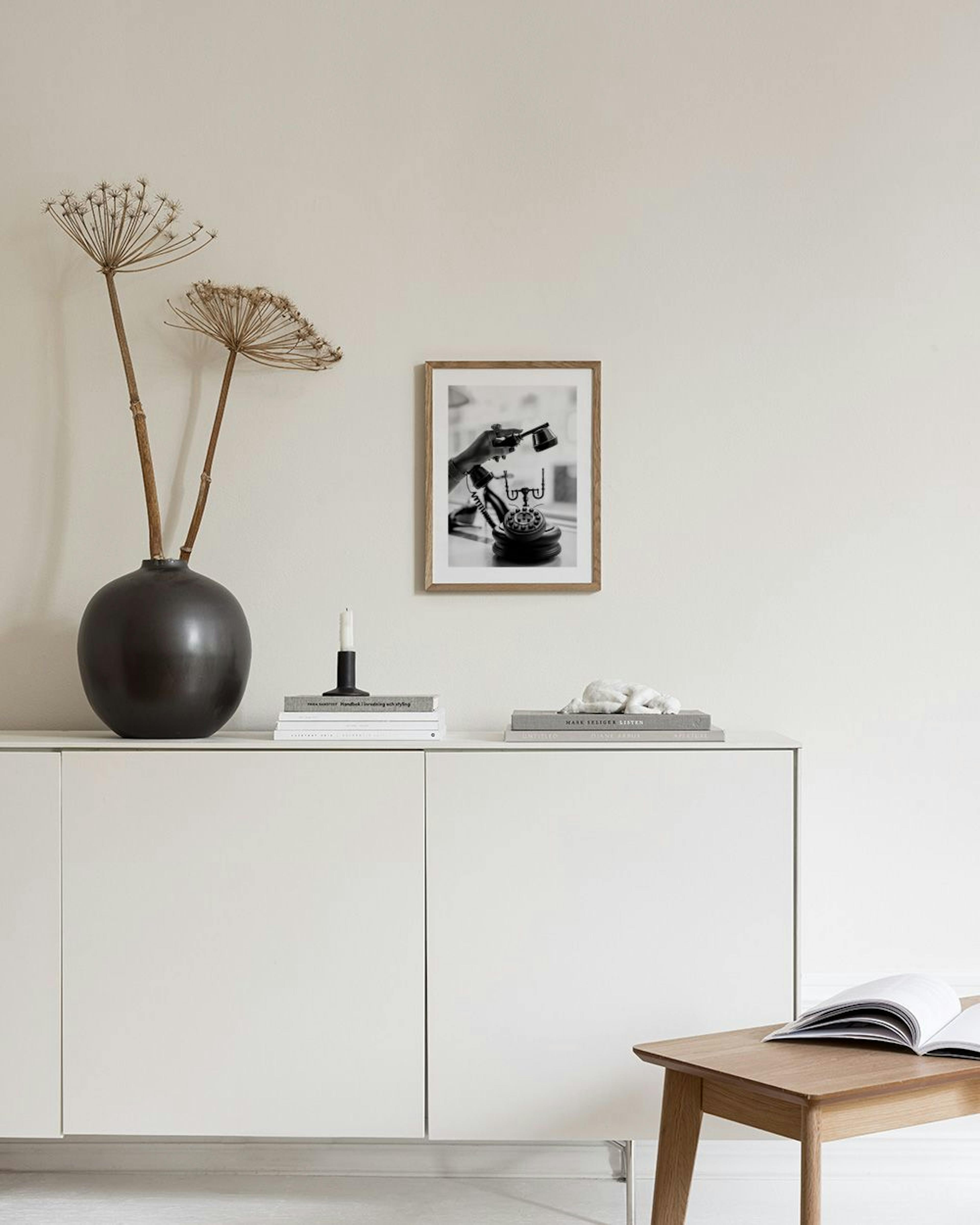 A black and white print of a hand holding a vintage telephone receiver, displayed on a grey cabinet with books and decor.
