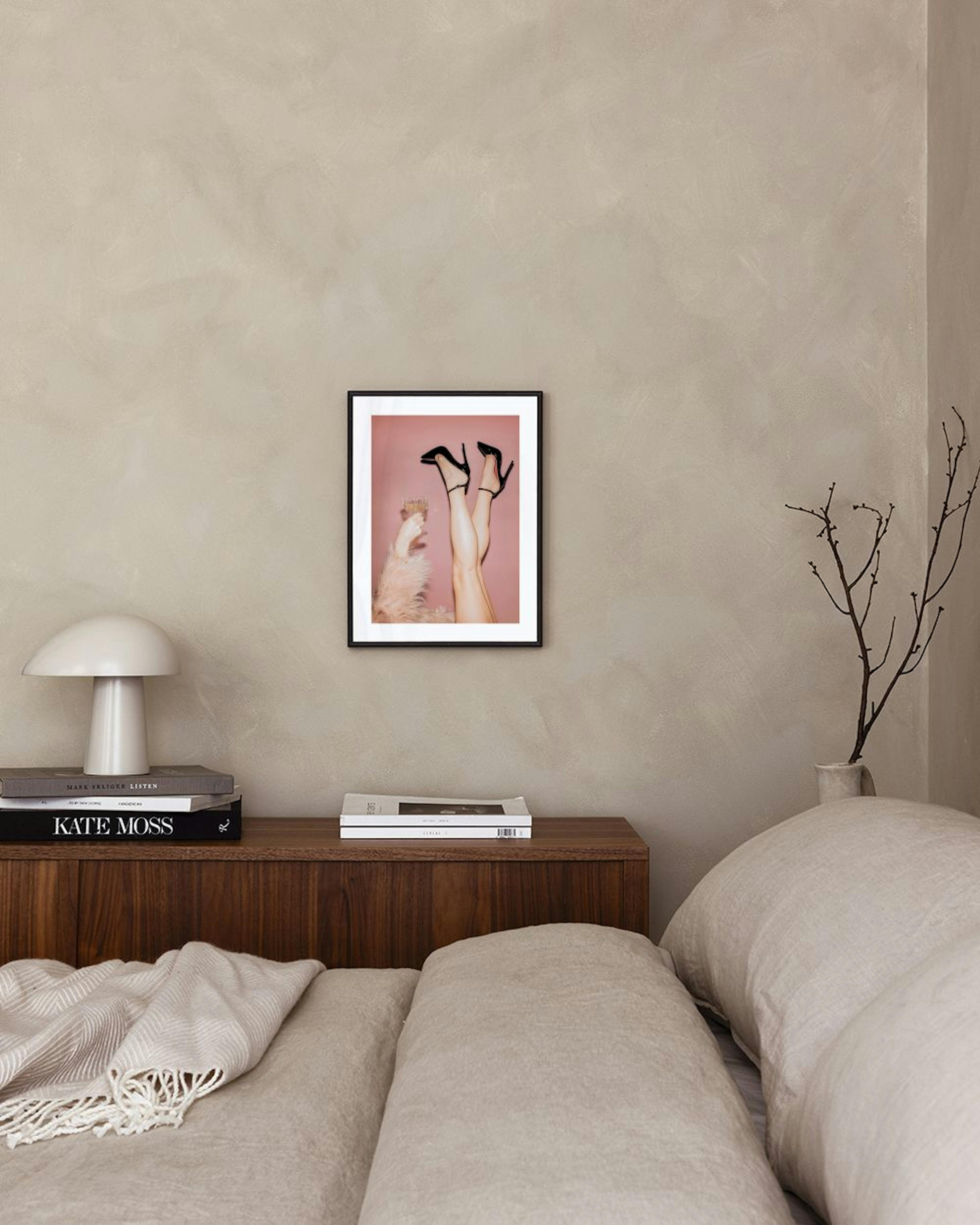 Poster met benen in zwarte hakken en een hand met champagneglas, hangend aan een roze muur boven een zwarte bijzettafel.