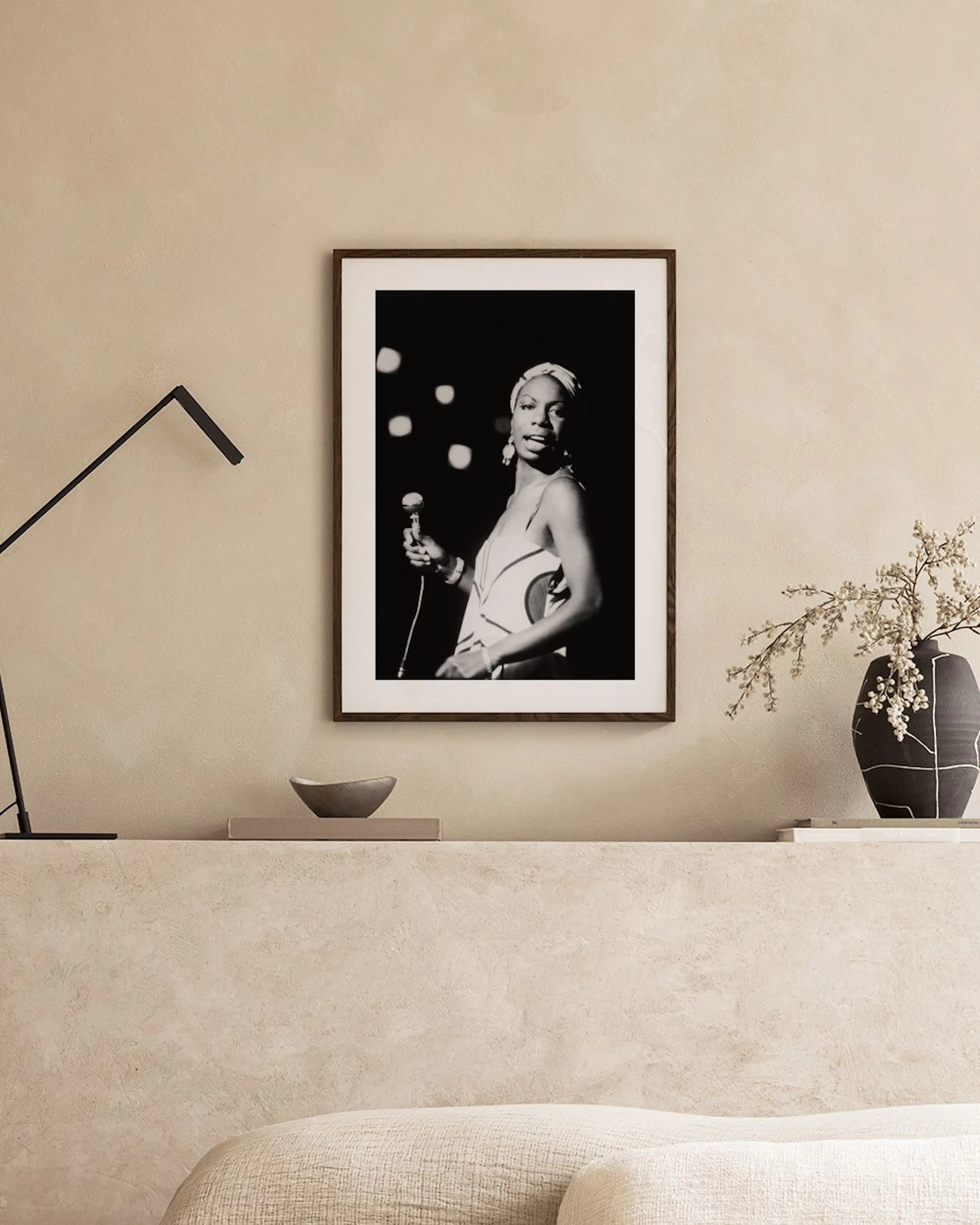 Svart-hvitt plakat av Nina Simone med mikrofon, stående på et konsollbord med lamper og dekor.