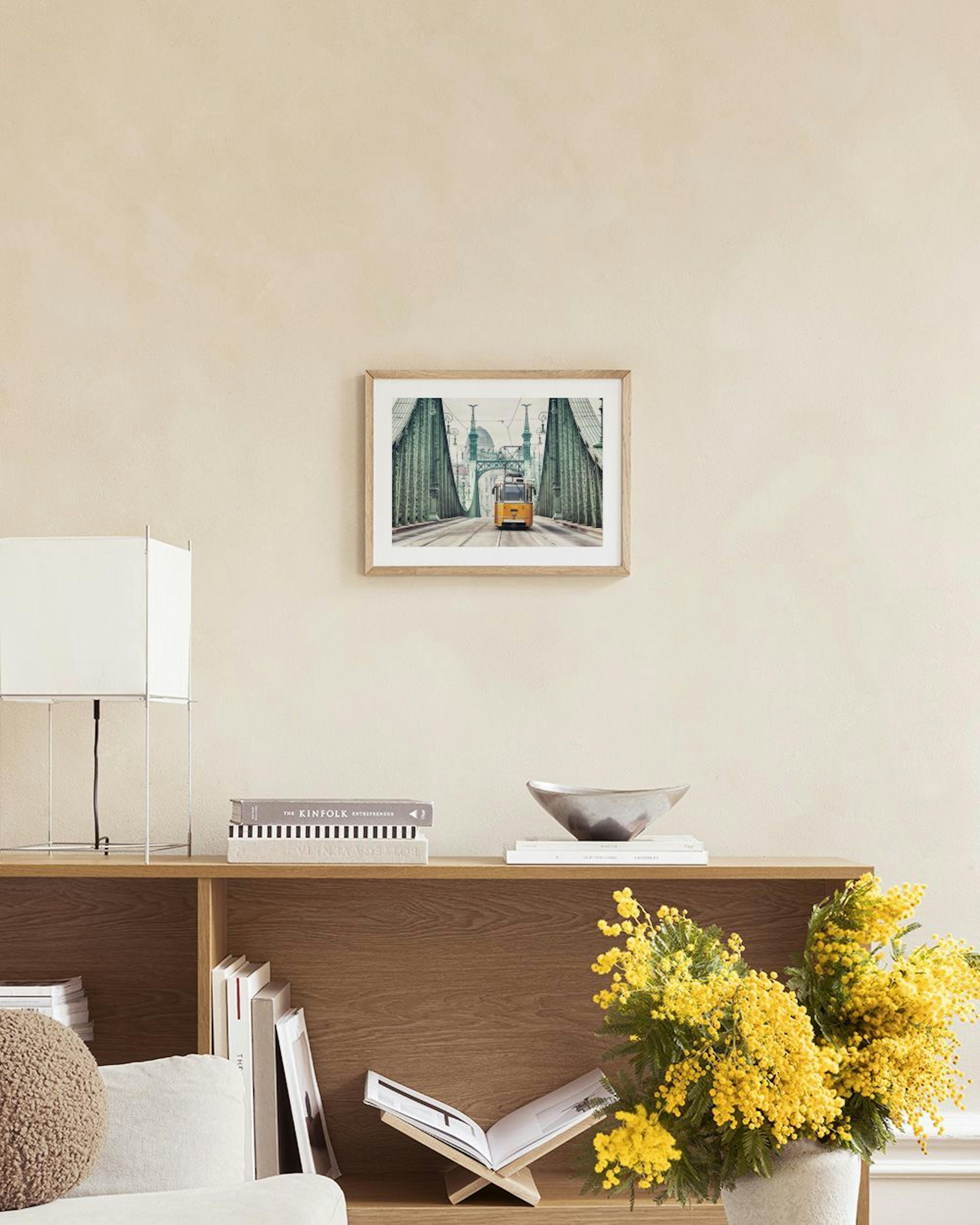 Poster einer gelben Straßenbahn auf einer grünen Brücke, über einer Bank mit Büchern und Vase.
