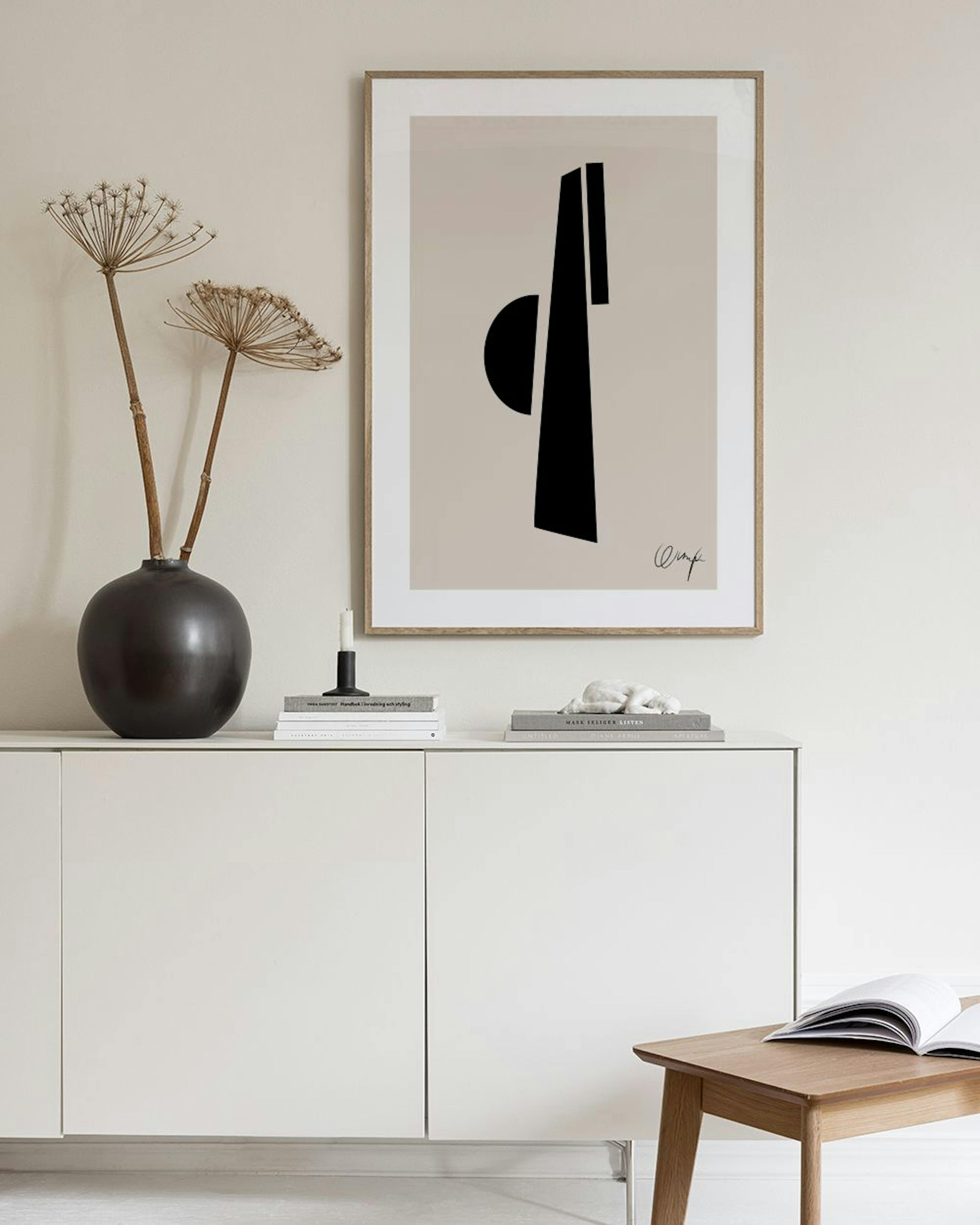 Plakat med svarte abstrakte former og en signatur, henger på en grå vegg ved siden av en vase med pampasgress og en stol.
