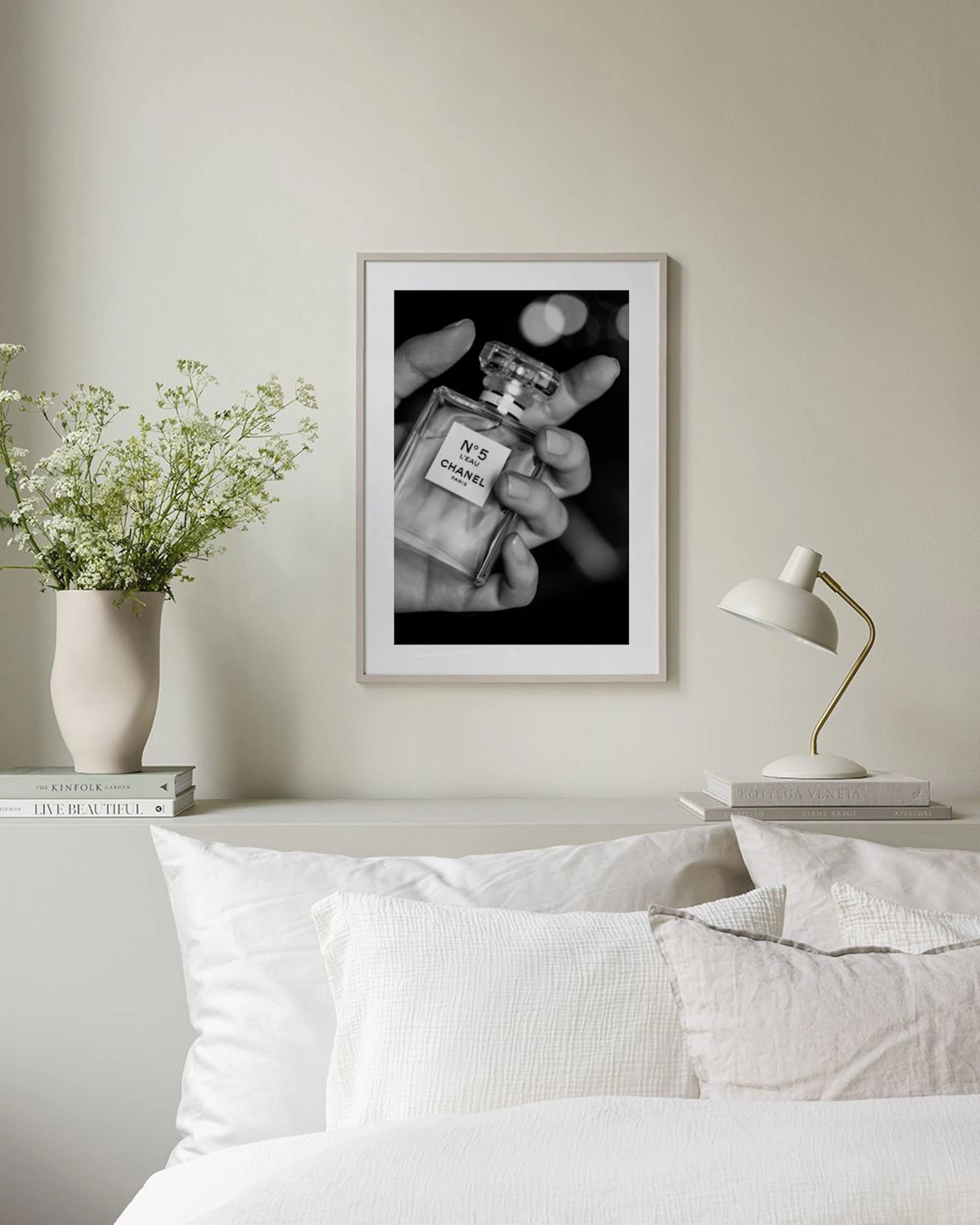 A black and white print of a hand holding a Chanel N°5 perfume bottle, displayed above a black shelf with books and decor in a m