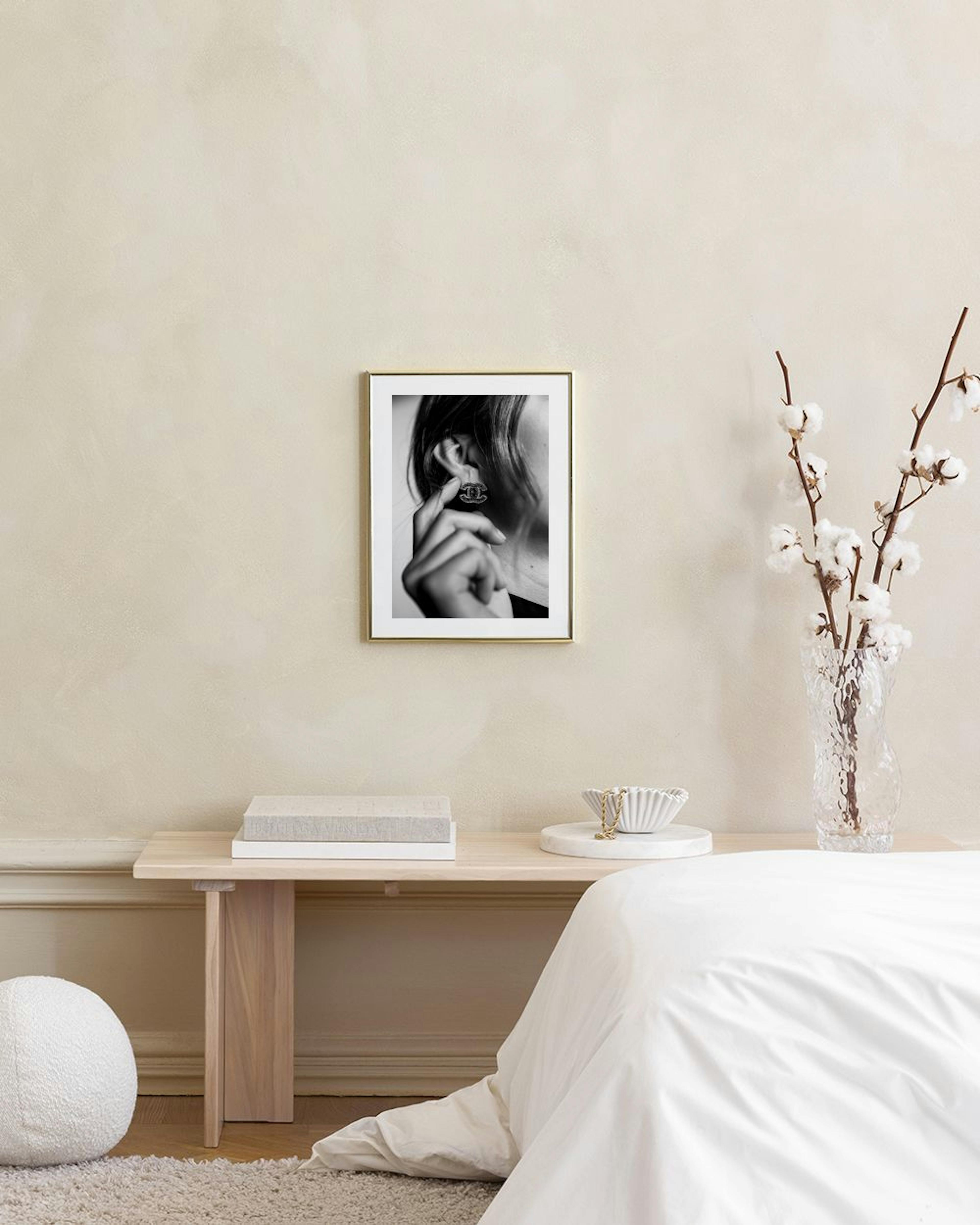 Póster de moda a preto e branco de uma mulher a colocar um brinco da Chanel, pendurado numa parede cinzenta acima de uma mesa de