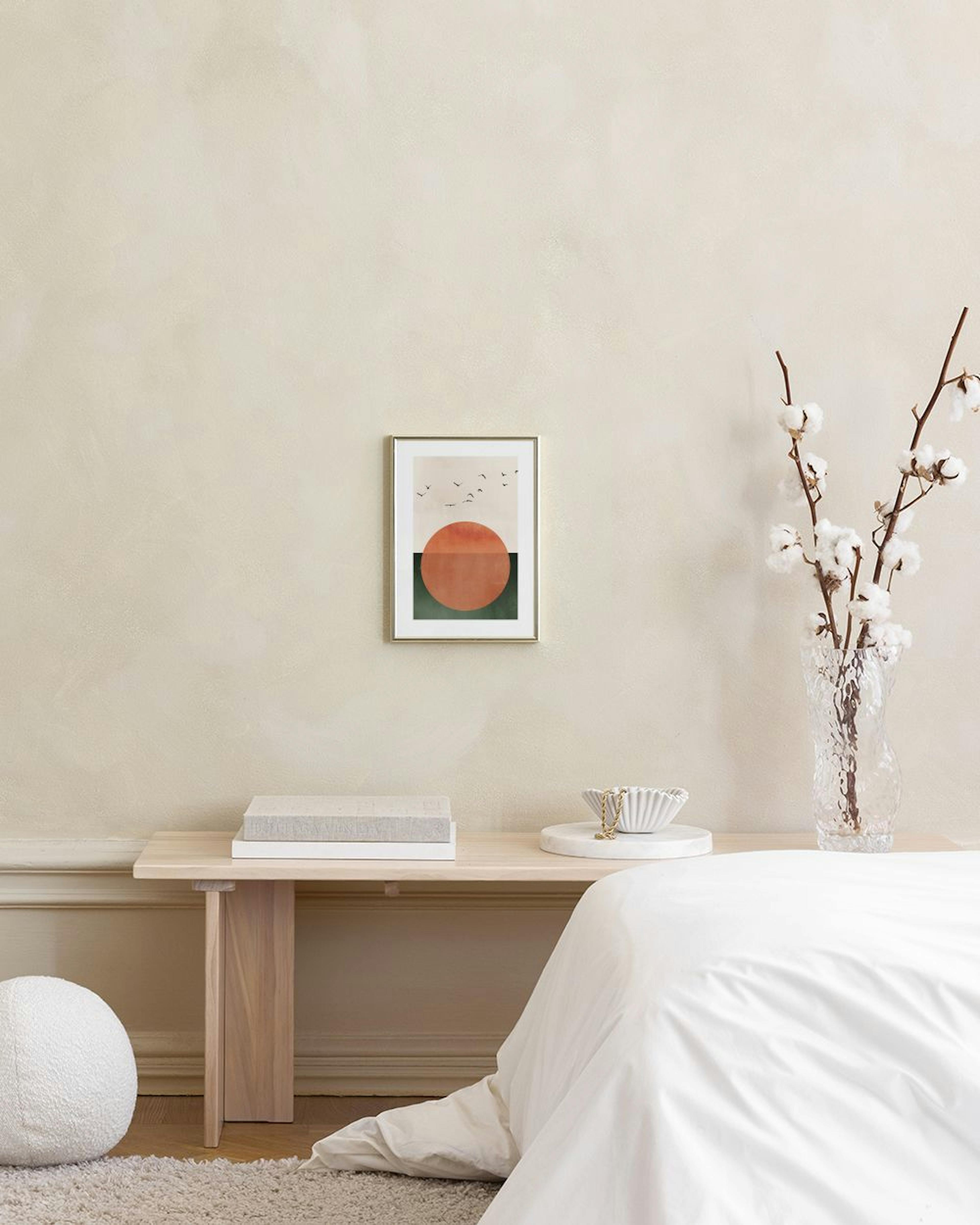 Póster de un sol naranja sobre un paisaje verde oscuro con pájaros volando, colgado sobre un mueble gris con libros y adornos.