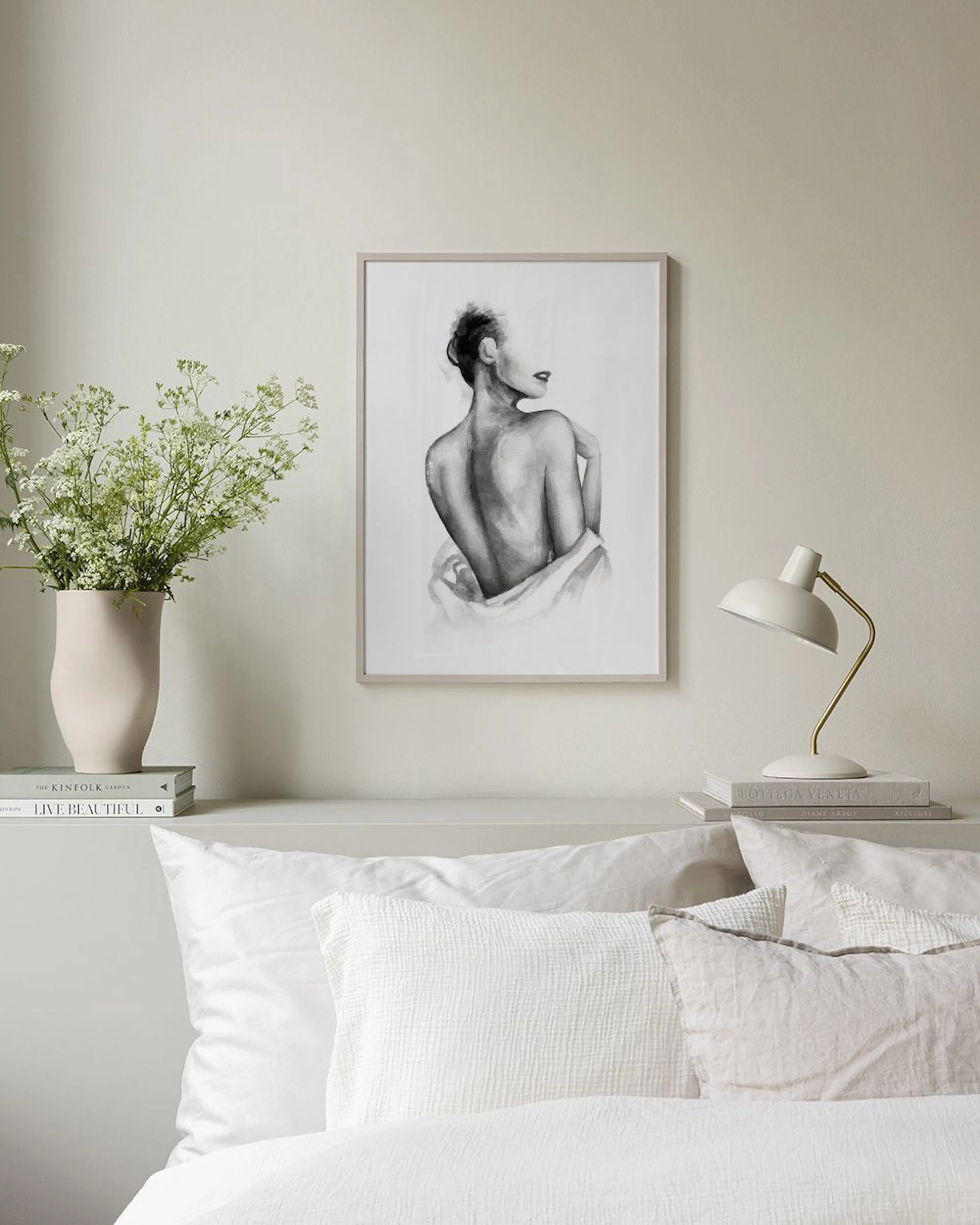 A black and white art print of a womans back, with dark red lips, hanging above a marble side table with white vases and books.