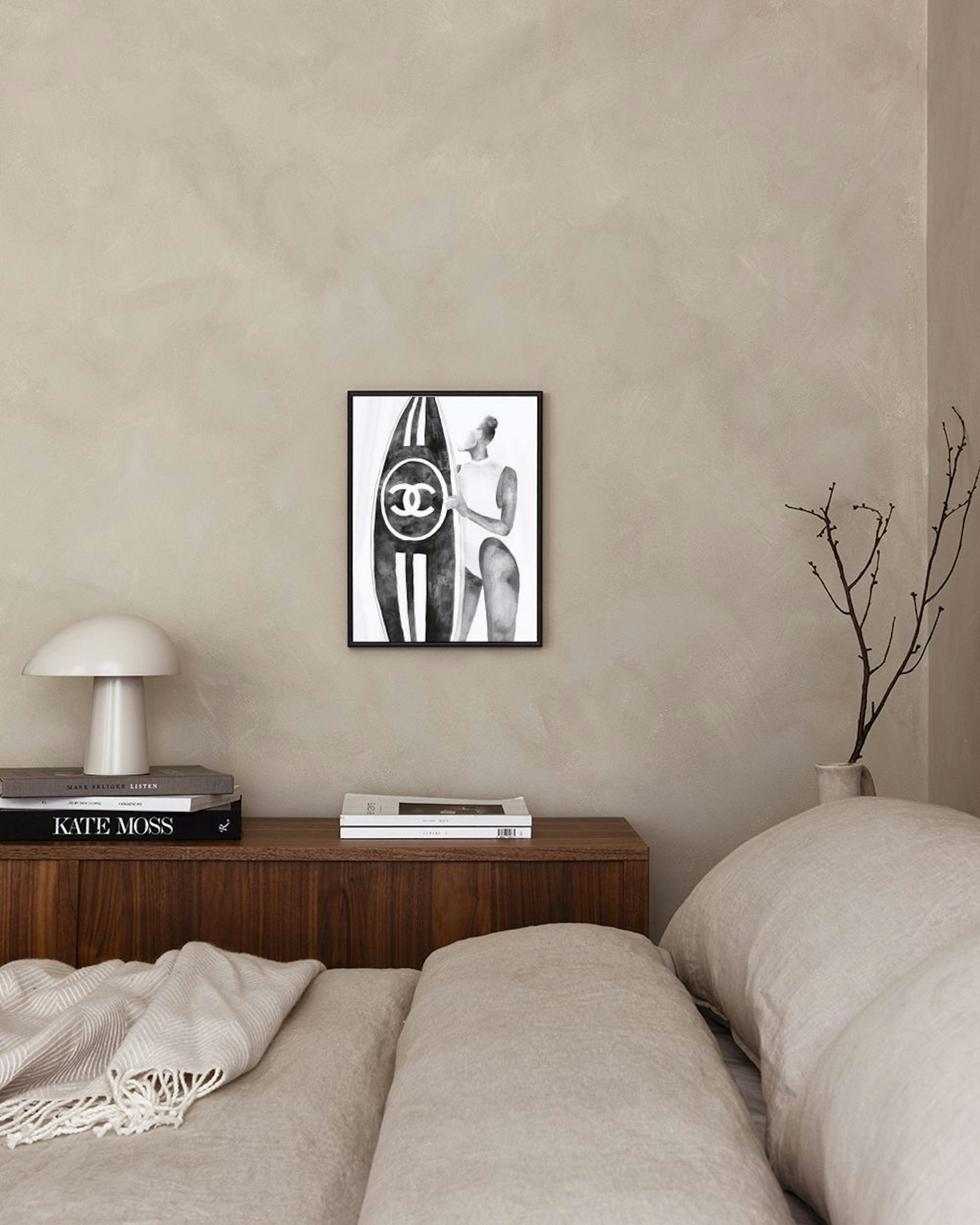 Black and white fashion print of a woman in a swimsuit holding a surfboard with a Chanel logo, displayed above a white cabinet.