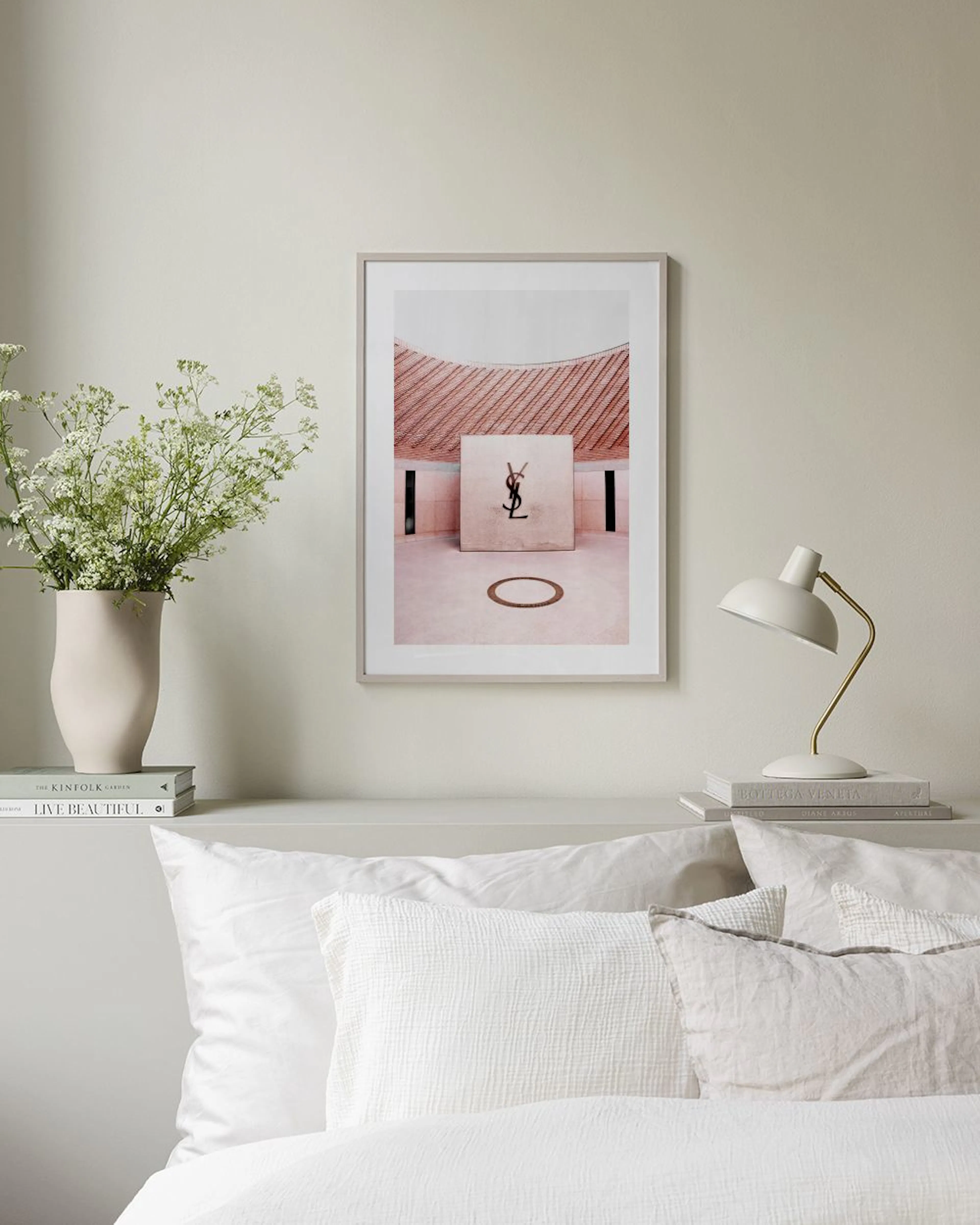 A print of a YSL beauty box with a gold circle on a pink-toned building, displayed above a beige cabinet with books and a lamp.