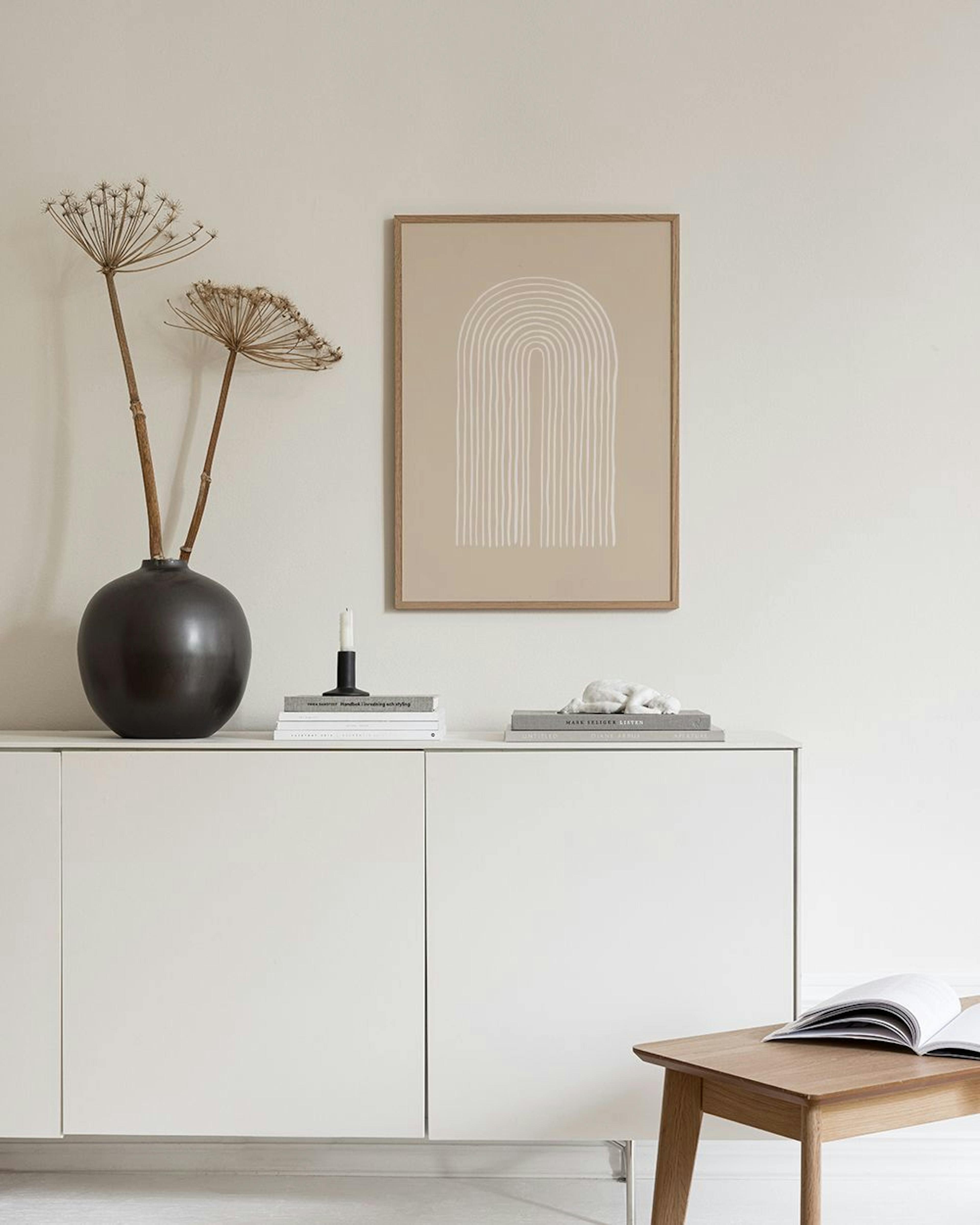 Un póster con un arco beige de líneas blancas, colgado en una pared gris sobre una mesa de mármol con jarrones y libros.