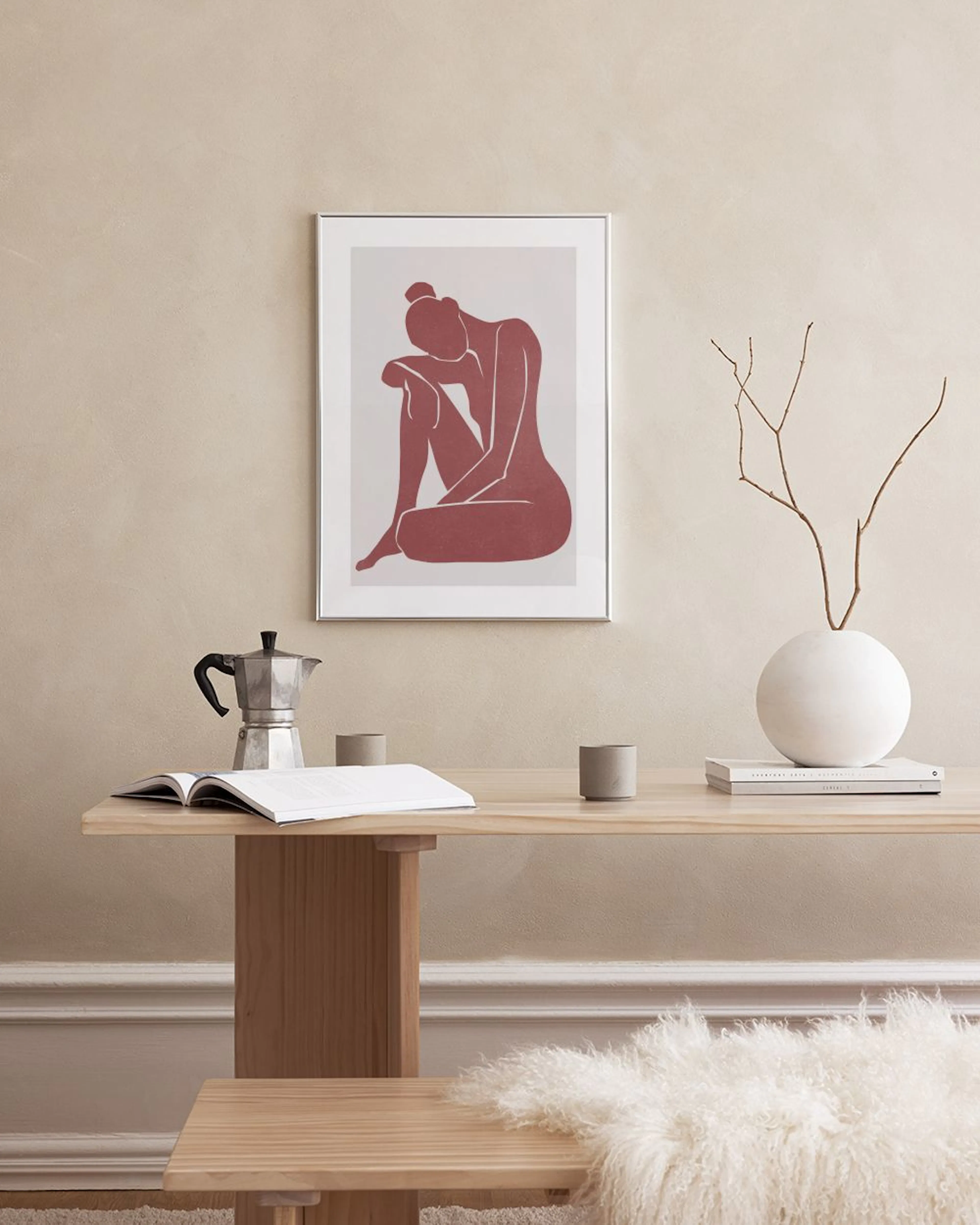 A print of a red female figure sitting with head resting on knees, displayed above a woven bench with decor.