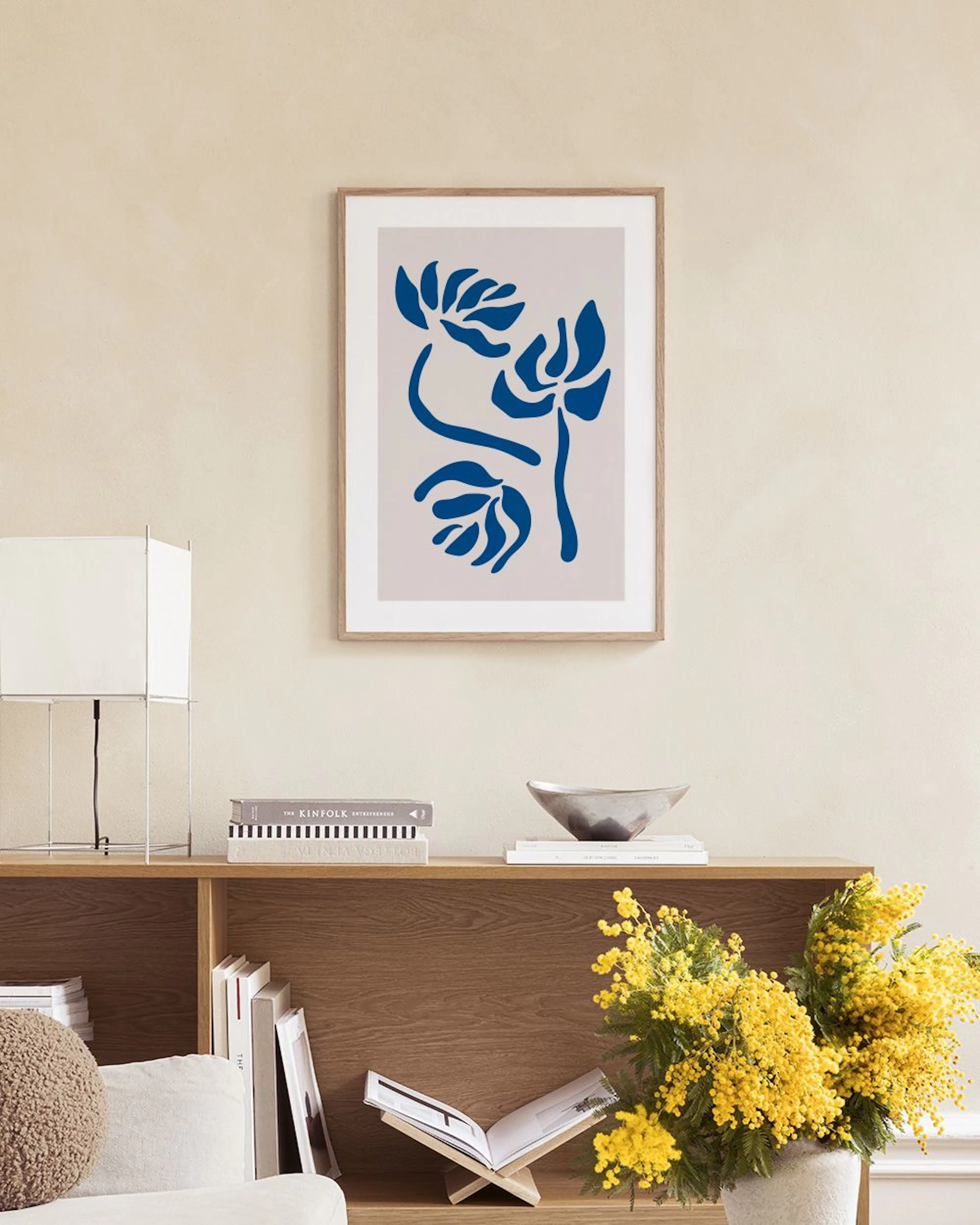 A print of three abstract blue floral shapes on a beige background, hanging in a light-colored room with a wooden chair and side