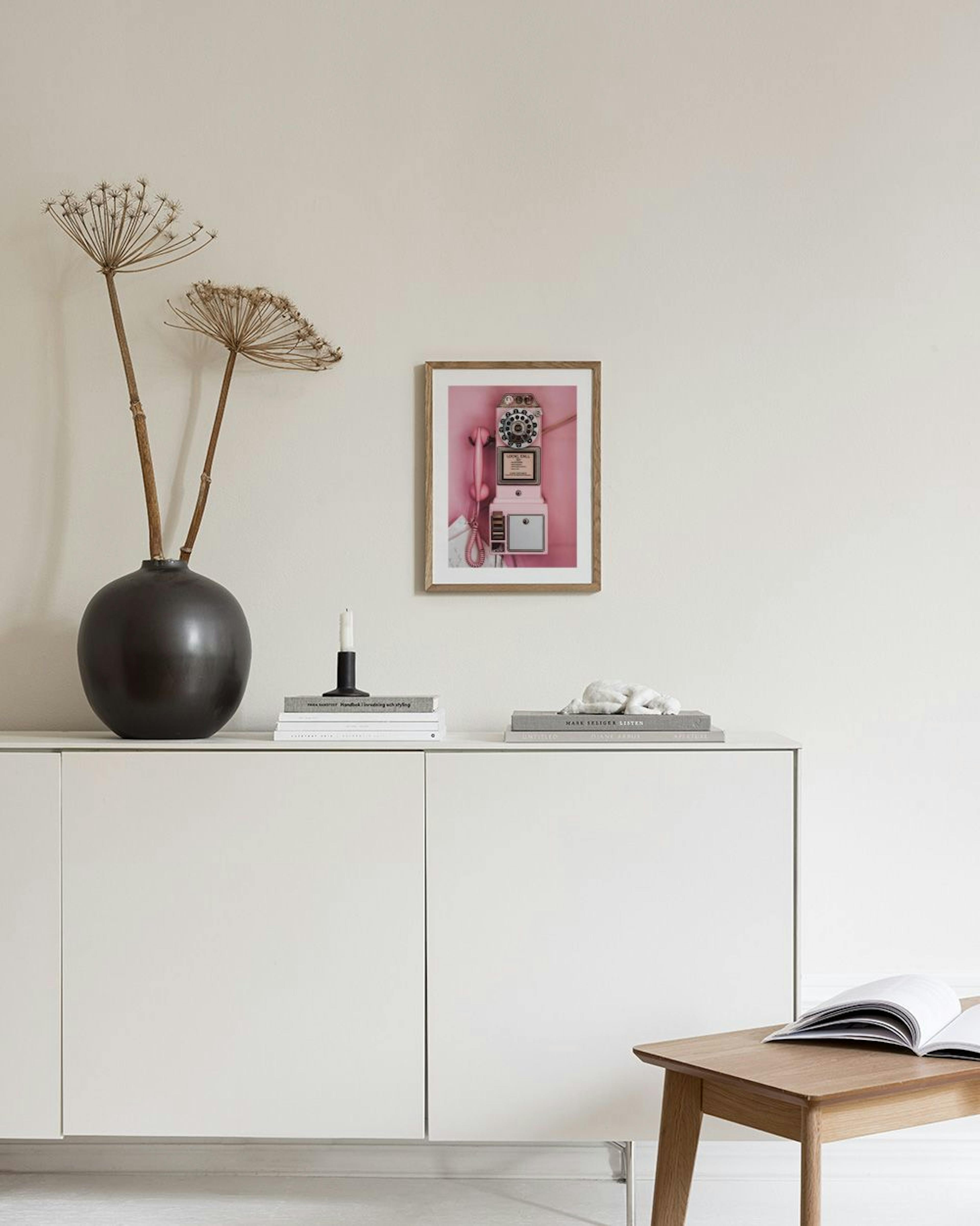 Póster de un teléfono de monedas rosa vintage en una pared gris, junto a un jarrón con flores y un puf rosa.