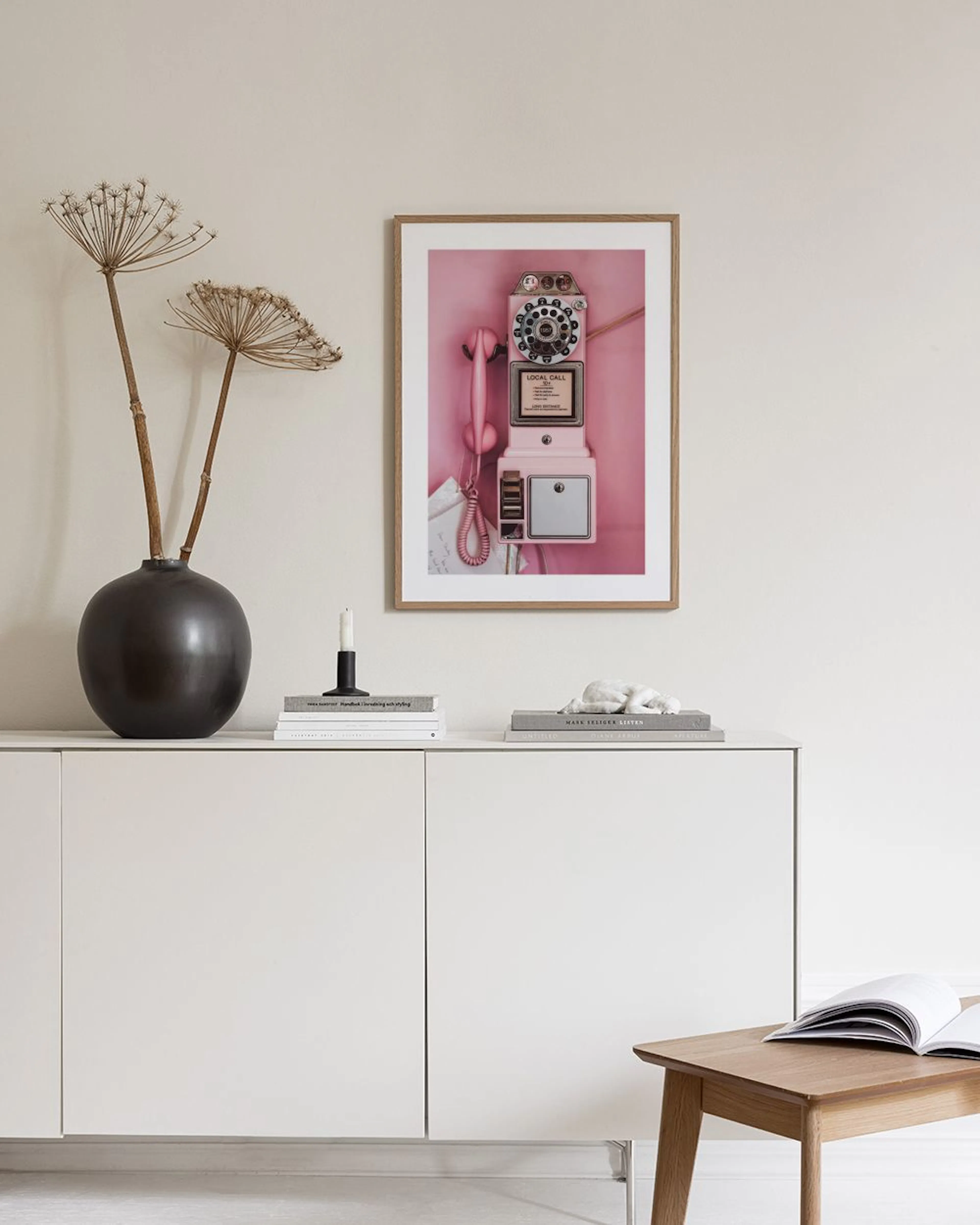 A print of a vintage pink payphone, hanging on a light grey wall next to a vase with flowers and a pink velvet ottoman.