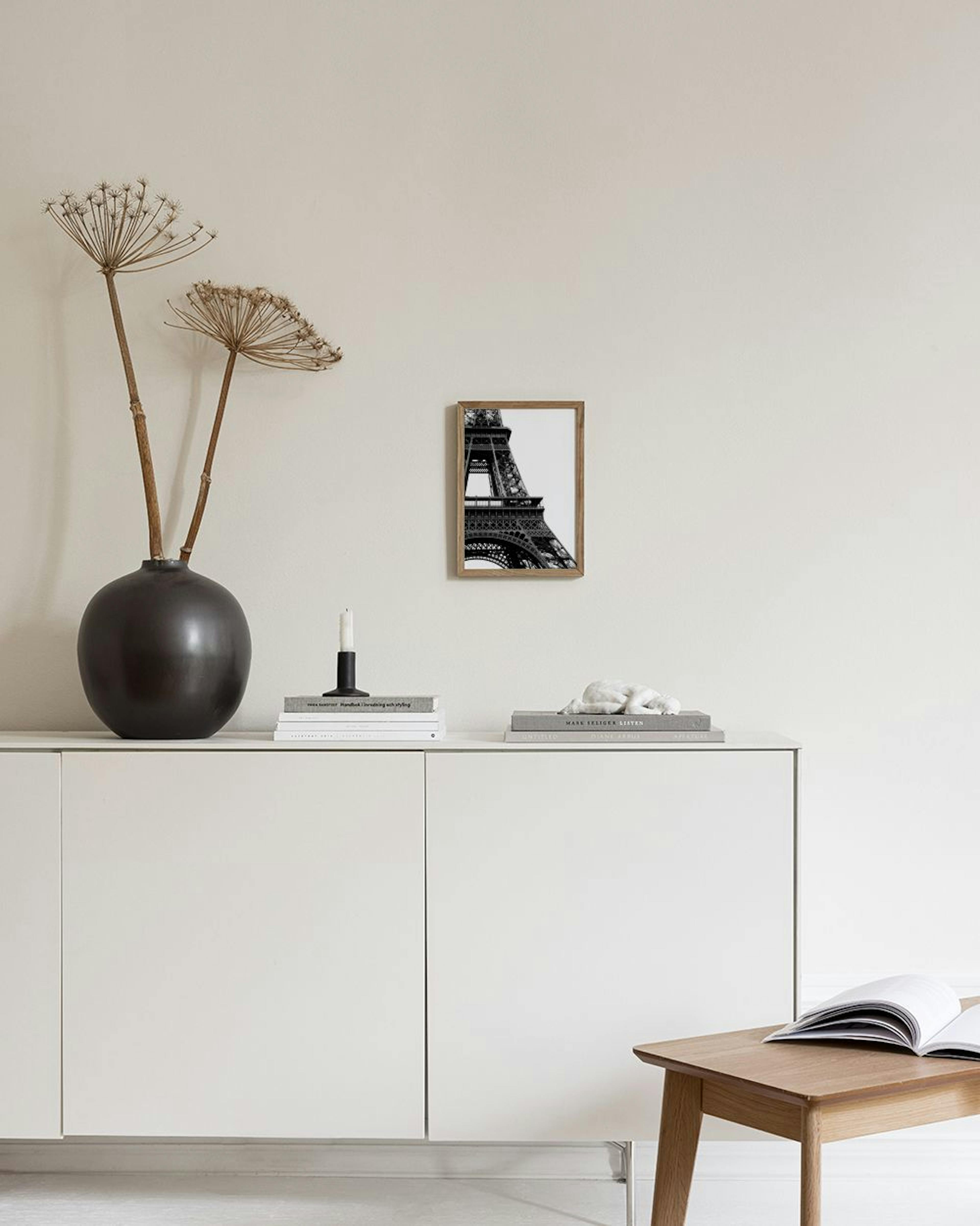 Zwart-wit poster van de Eiffeltoren, hangend boven een donkere houten kast met boeken en een witte schaal.