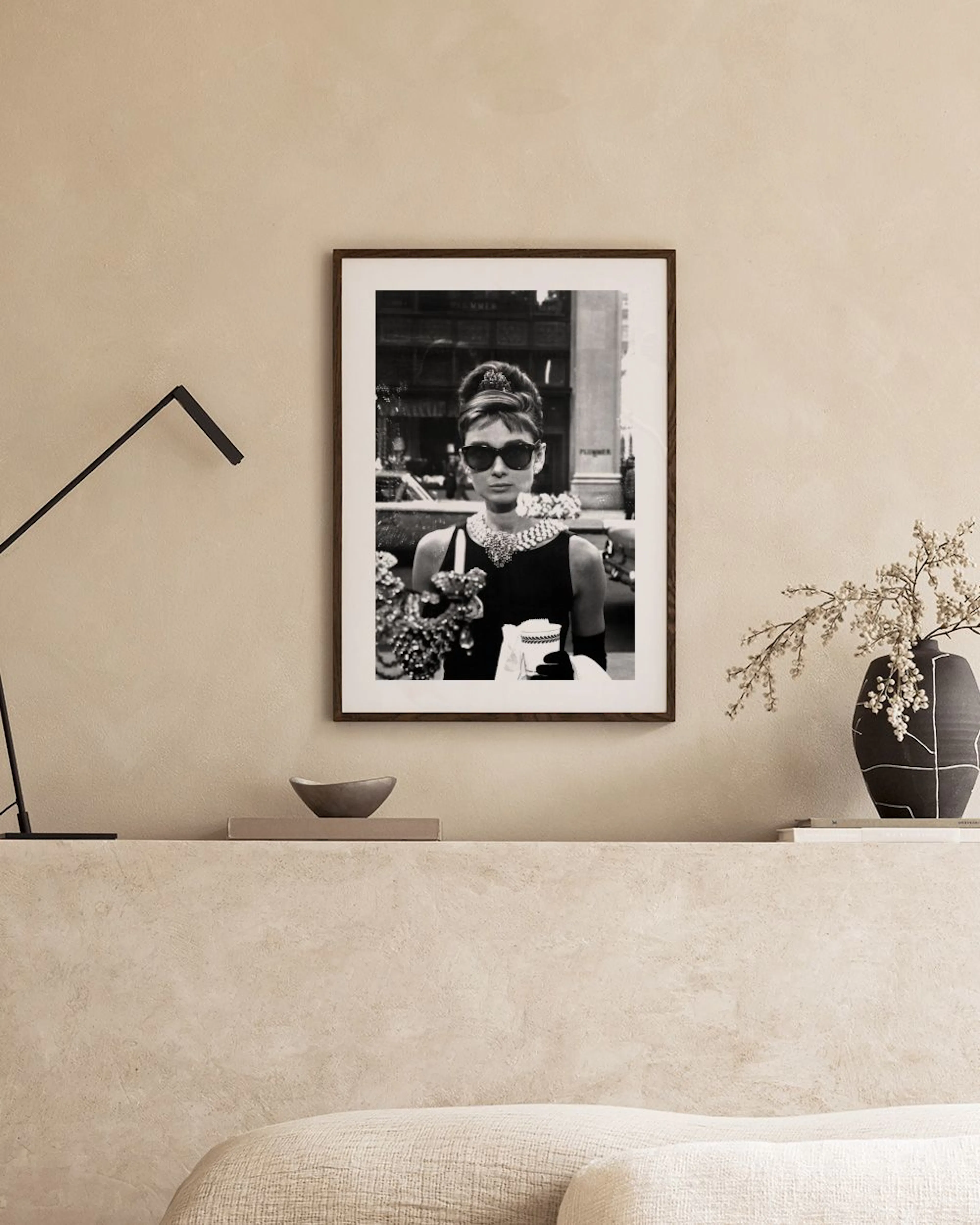 Czarno-biały plakat z Audrey Hepburn w okularach przeciwsłonecznych, wiszący nad betonową ławą. Obok wazon i miska.