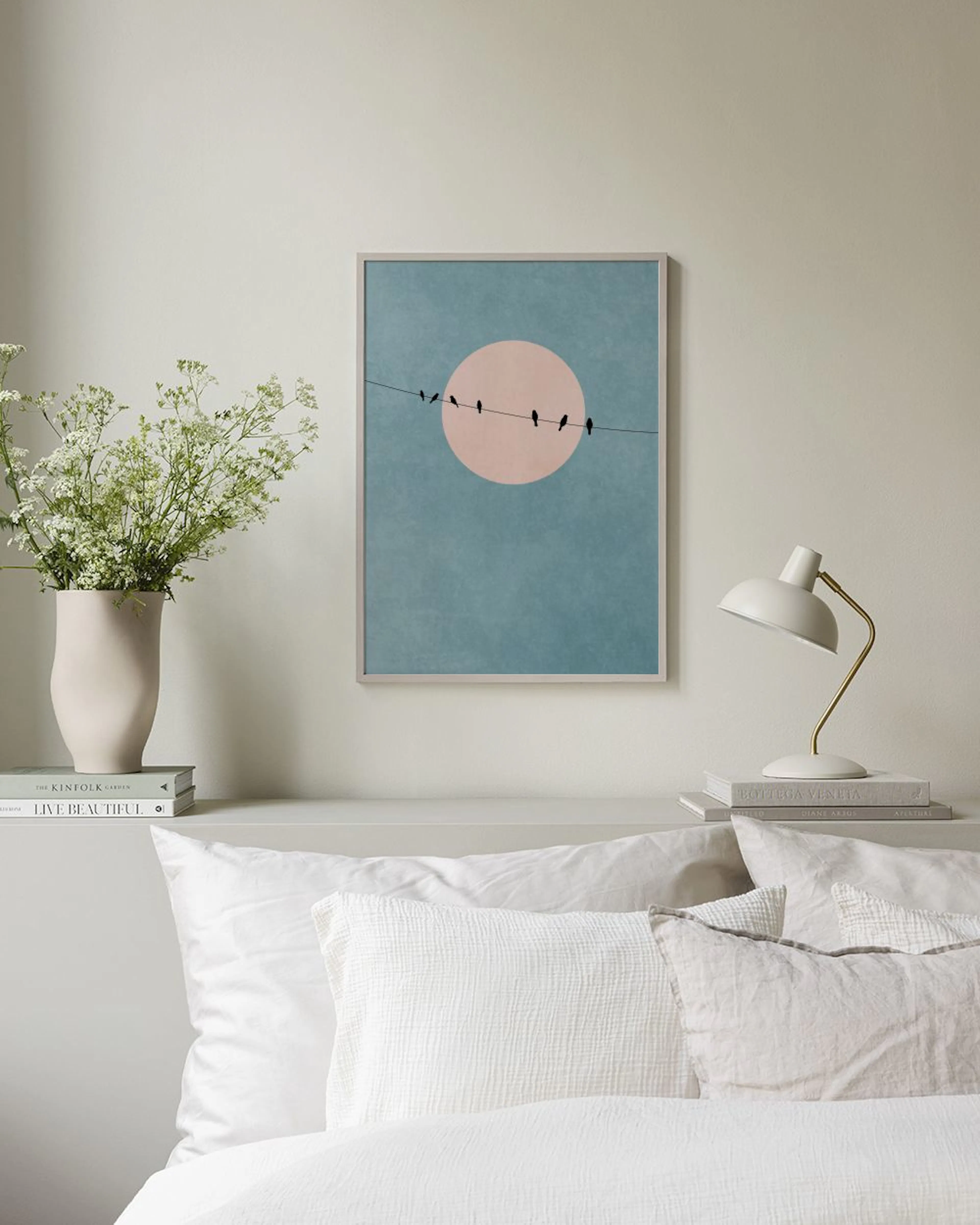 Poster minimalista com pássaros num fio, sol pêssego e fundo azul-claro, pendurado acima de um móvel branco com vaso.