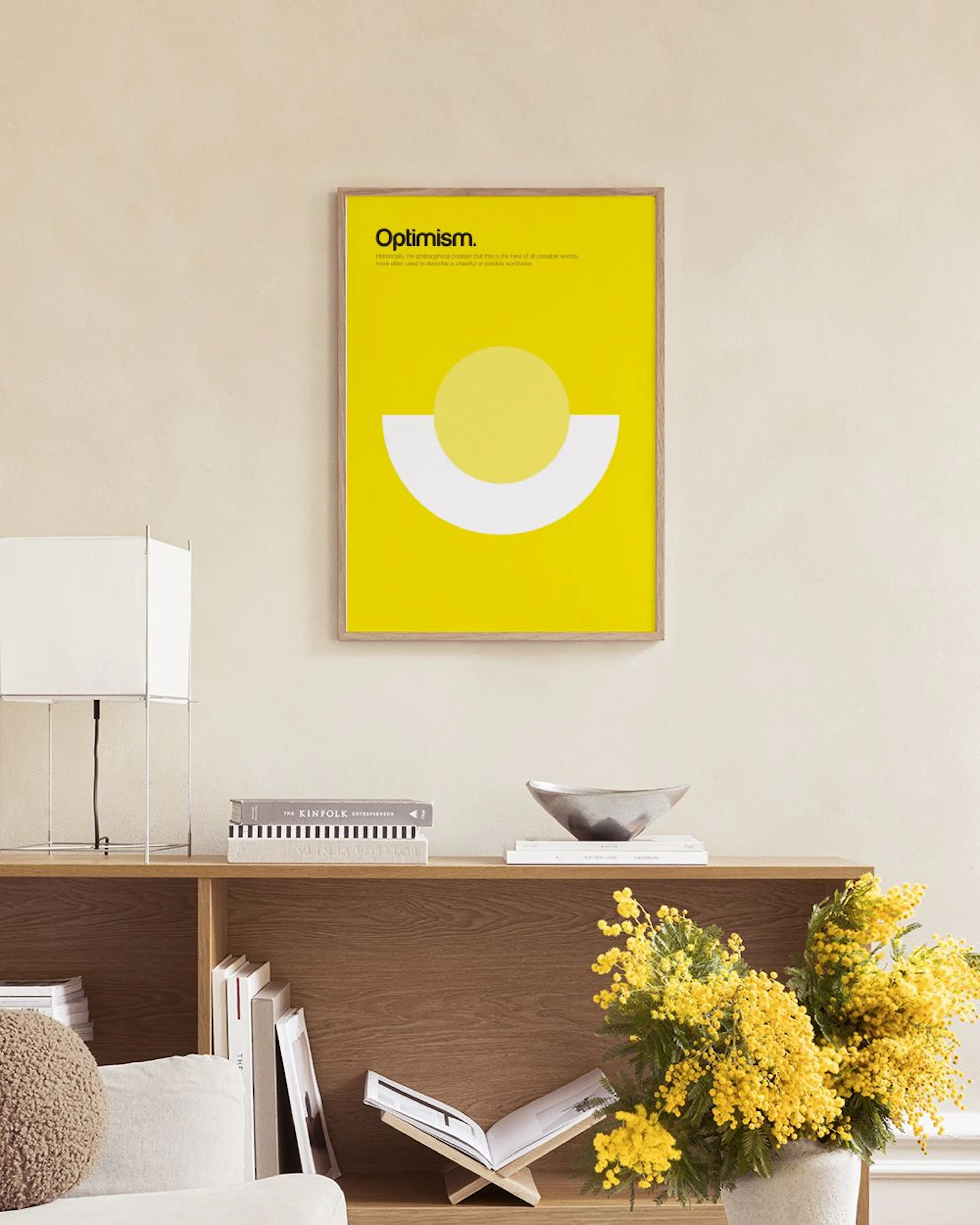 A bright yellow Optimism print with a light yellow circle and white semi-circle, hanging above a woven bench with books.
