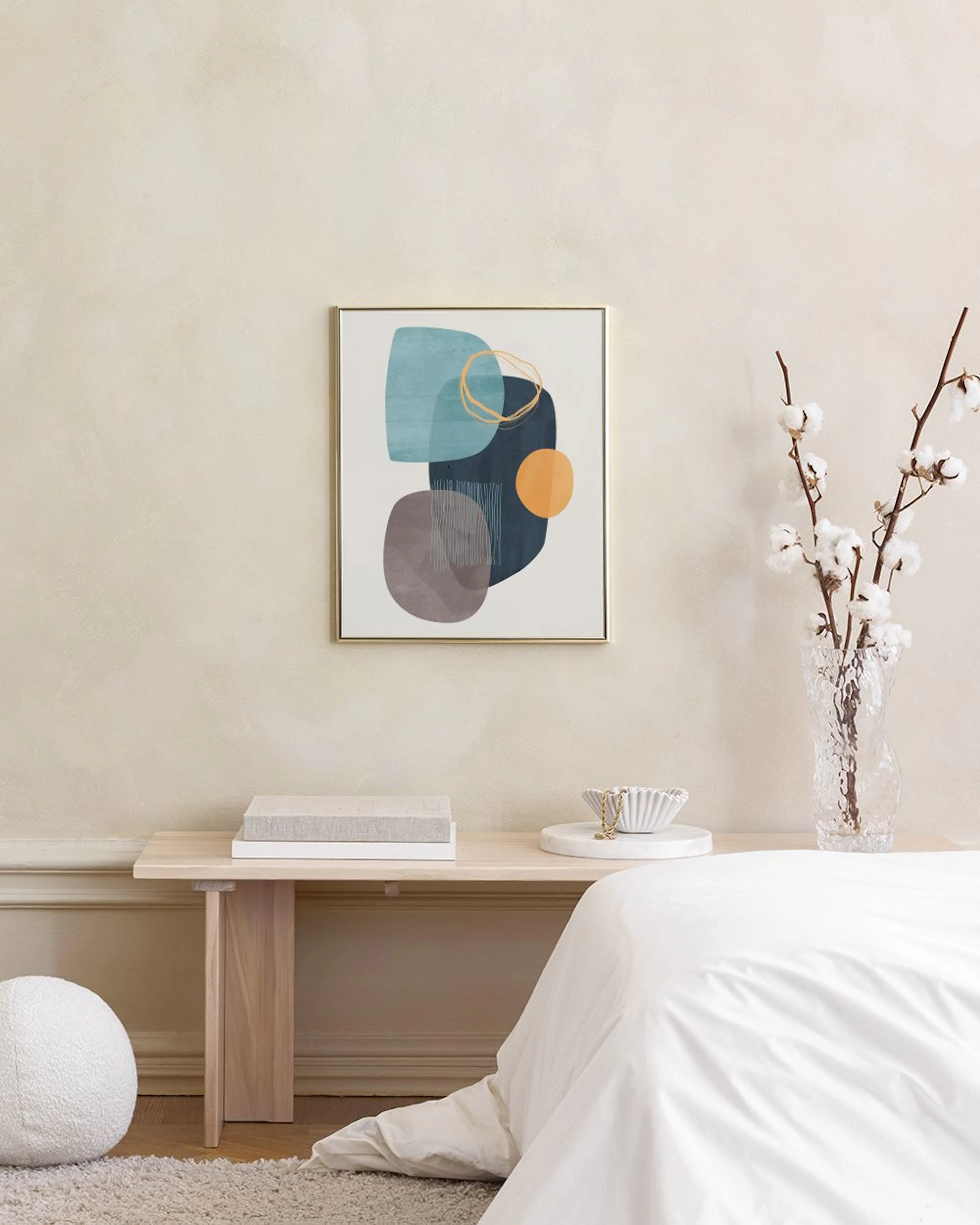 A print of abstract shapes in teal, dark blue, and grey with an orange circle, hanging above a white cabinet with books and a va