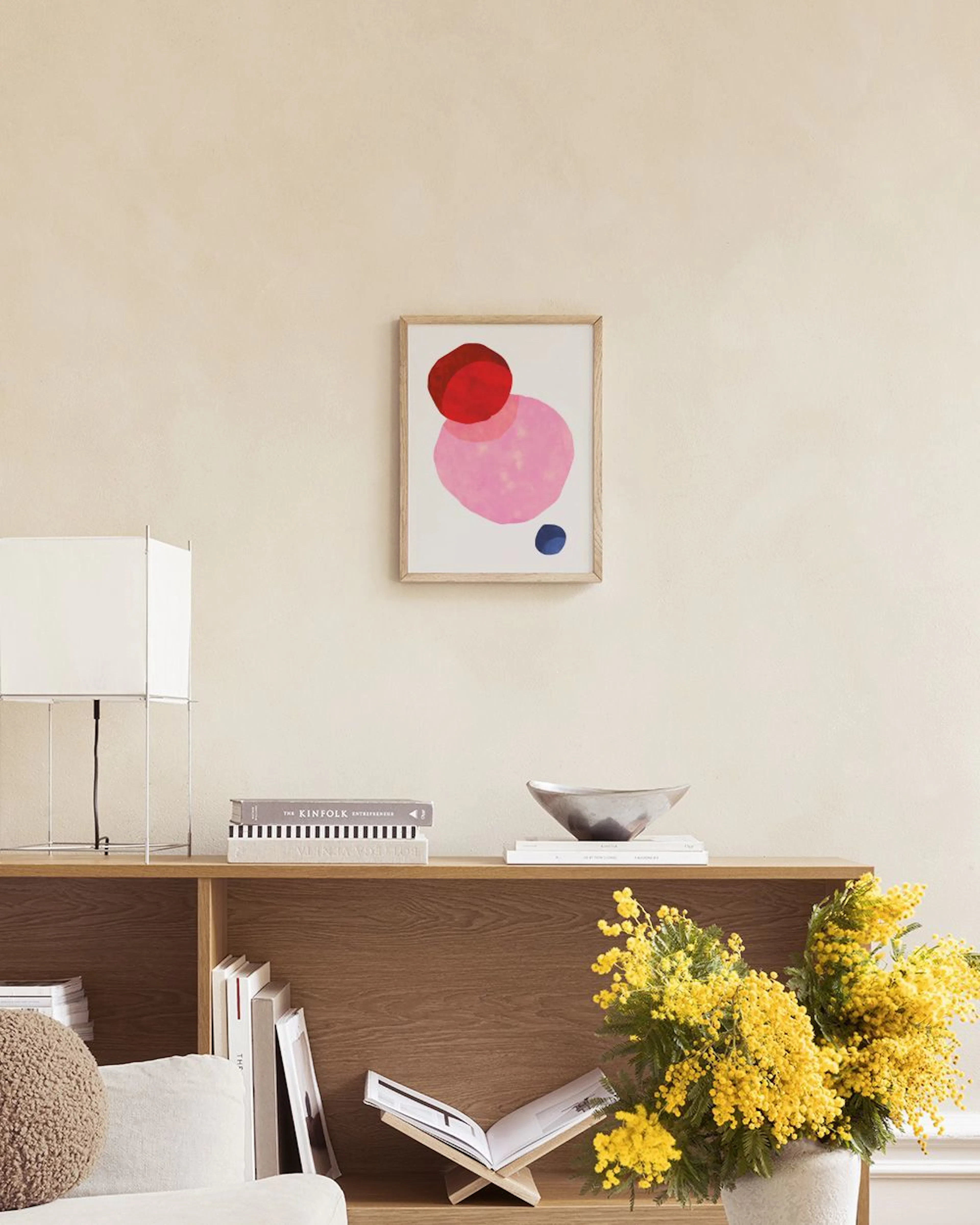 Poster con forme astratte rosa, rosse e blu, appeso sopra uno sgabello con libri in una stanza moderna.