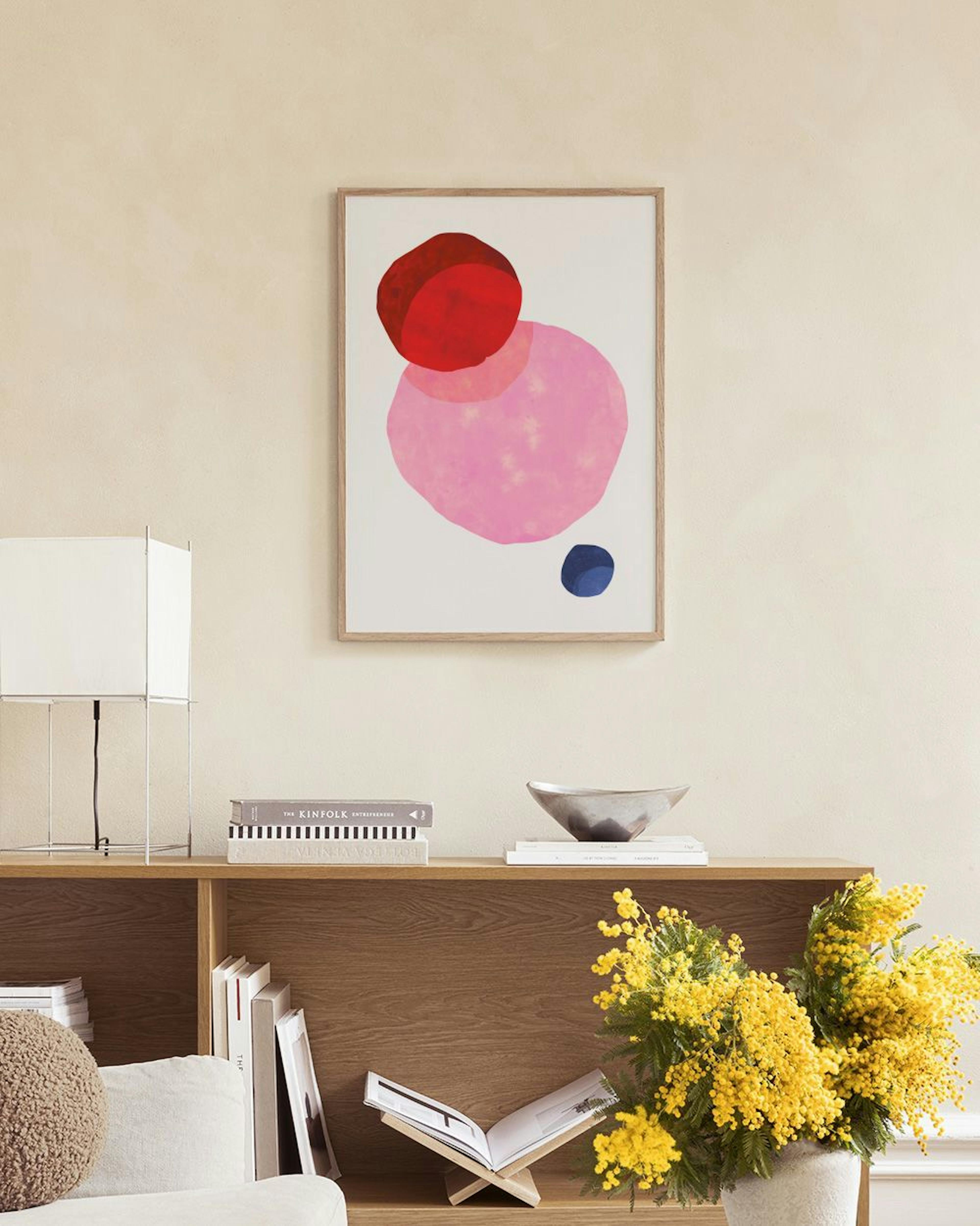 Poster con forme astratte rosso, rosa e blu, appeso sopra uno sgabello di sughero con libri in un ambiente luminoso.