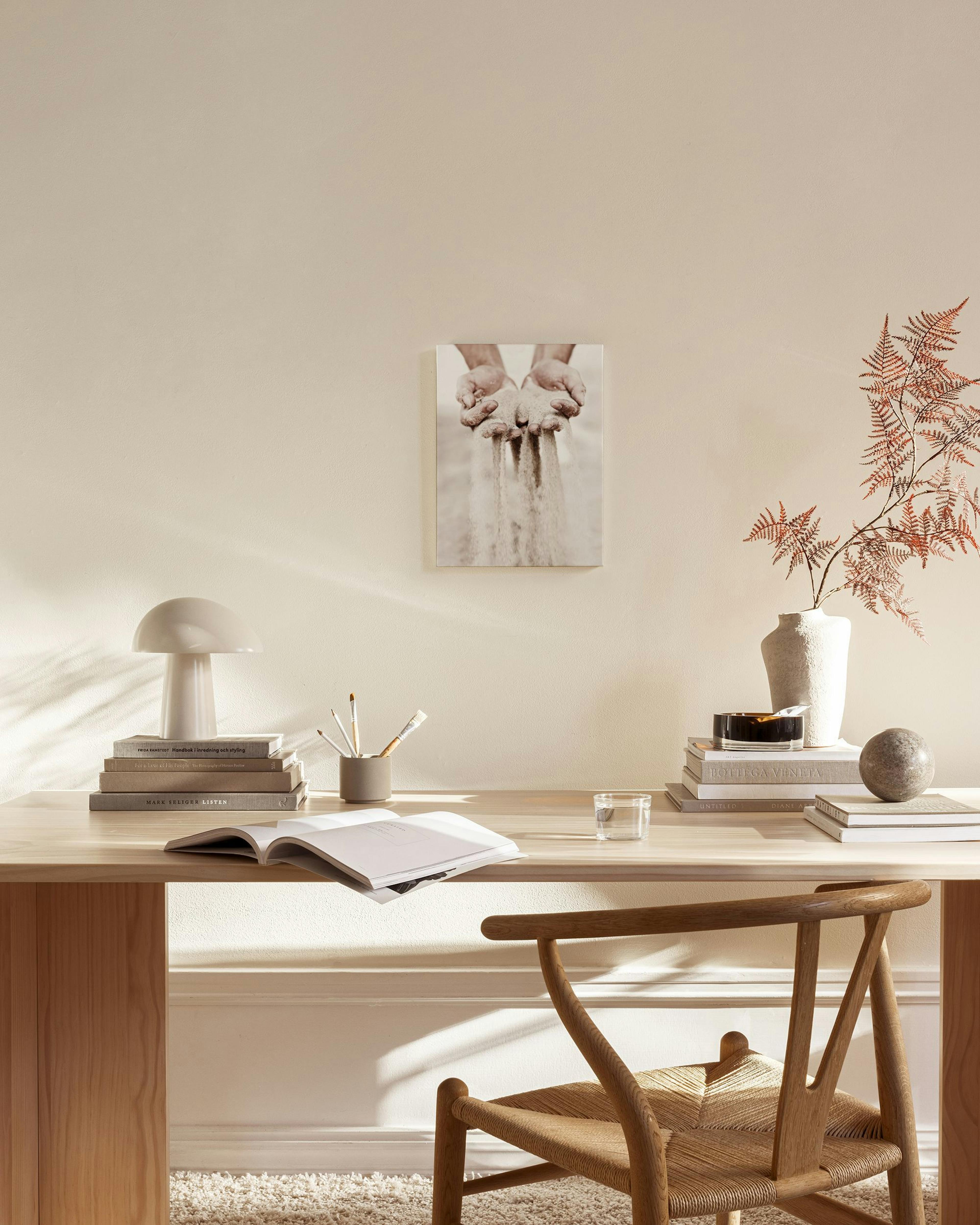 Canvas met handen die zand laten vallen, hangend in een lichte, moderne woonkamer met een houten bankje en stoel.