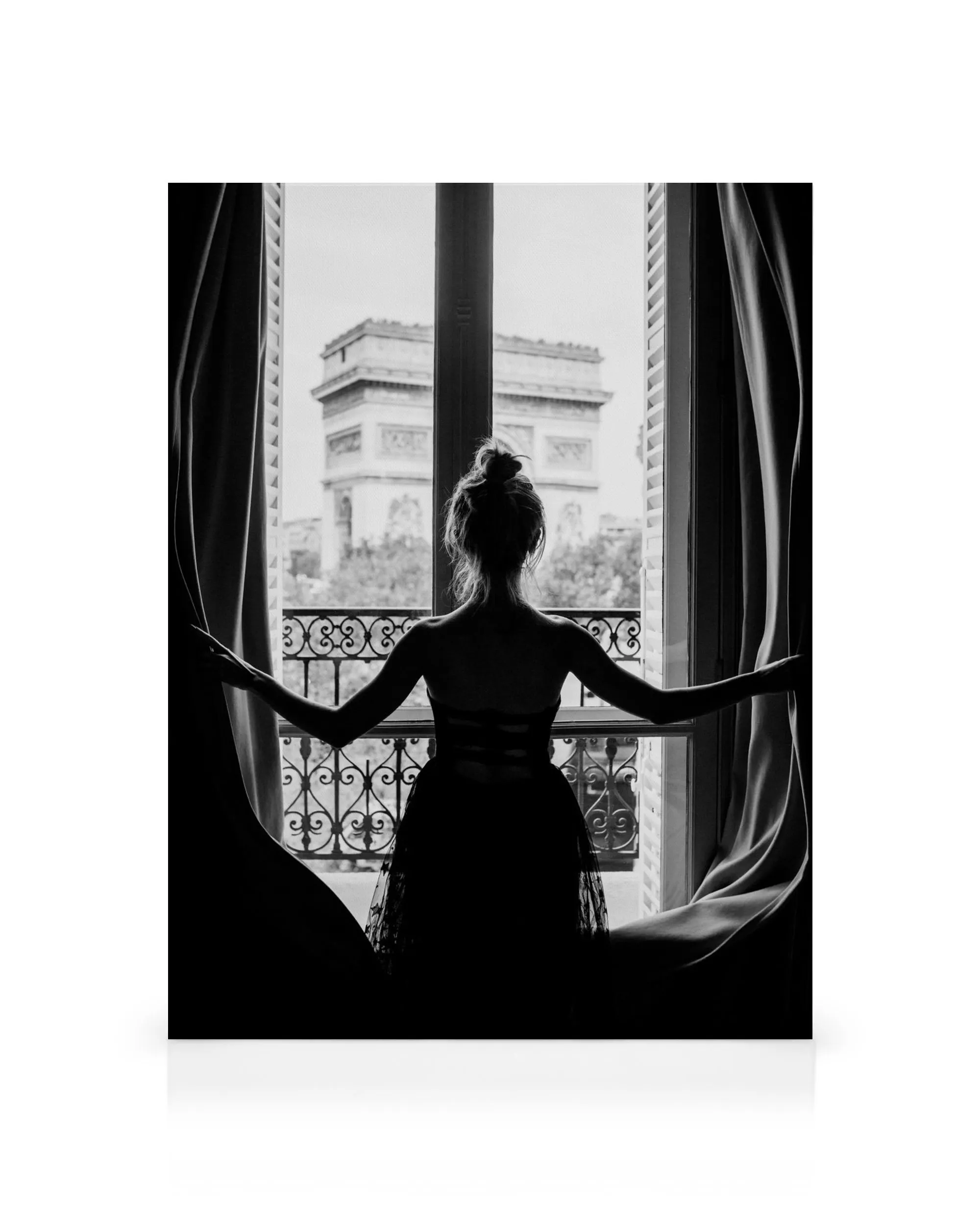 Black and white canvas print of a woman opening curtains, looking at a Paris arch monument from a window balcony.