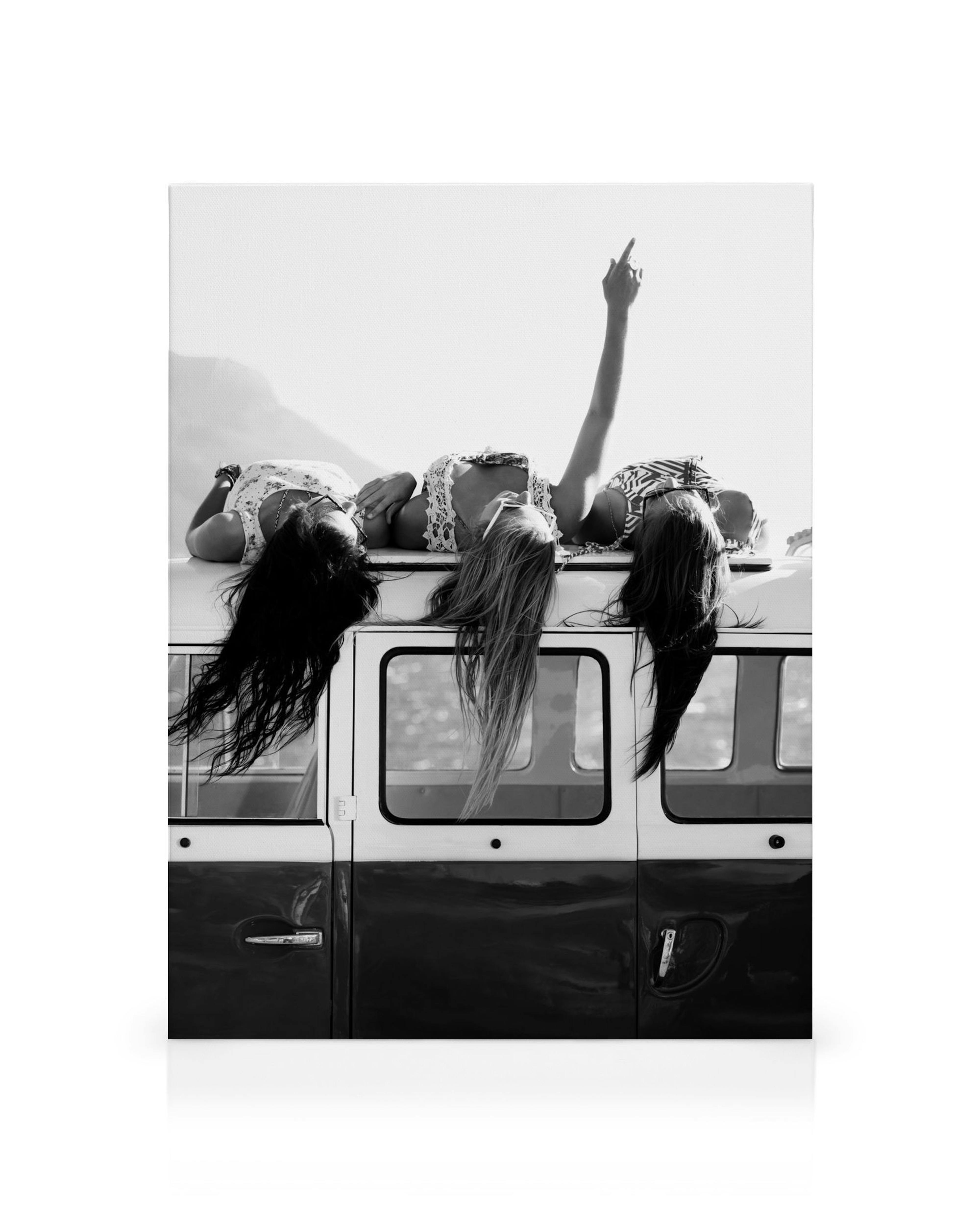 Zwart-wit canvas van drie vrouwen die op een busje liggen met wapperend haar, met uitzicht op bergen en water.